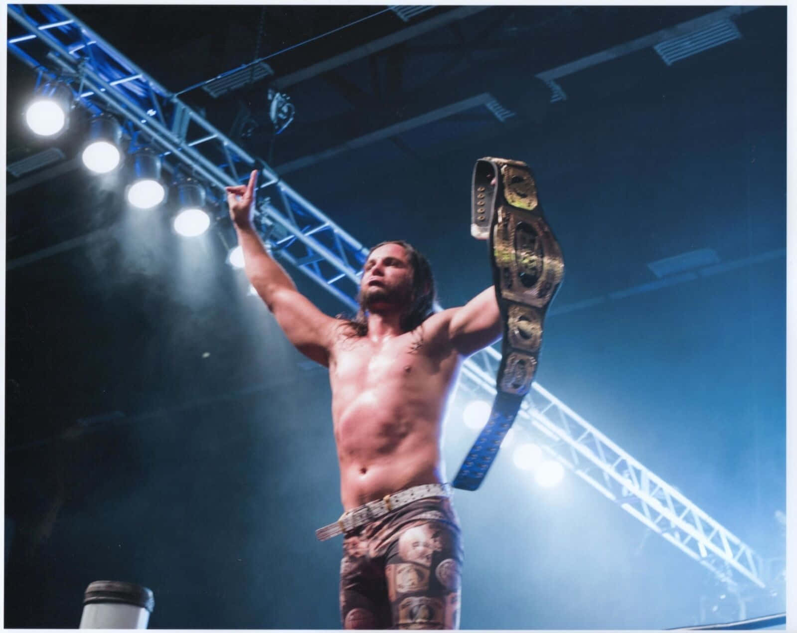 American Professional Wrestler Matt Jackson ROH Championship Belt 壁紙 — 一位赤裸上身的長髮摔角手在黑暗的競技場中，在明亮的舞台燈光下勝利地高舉冠軍腰帶。