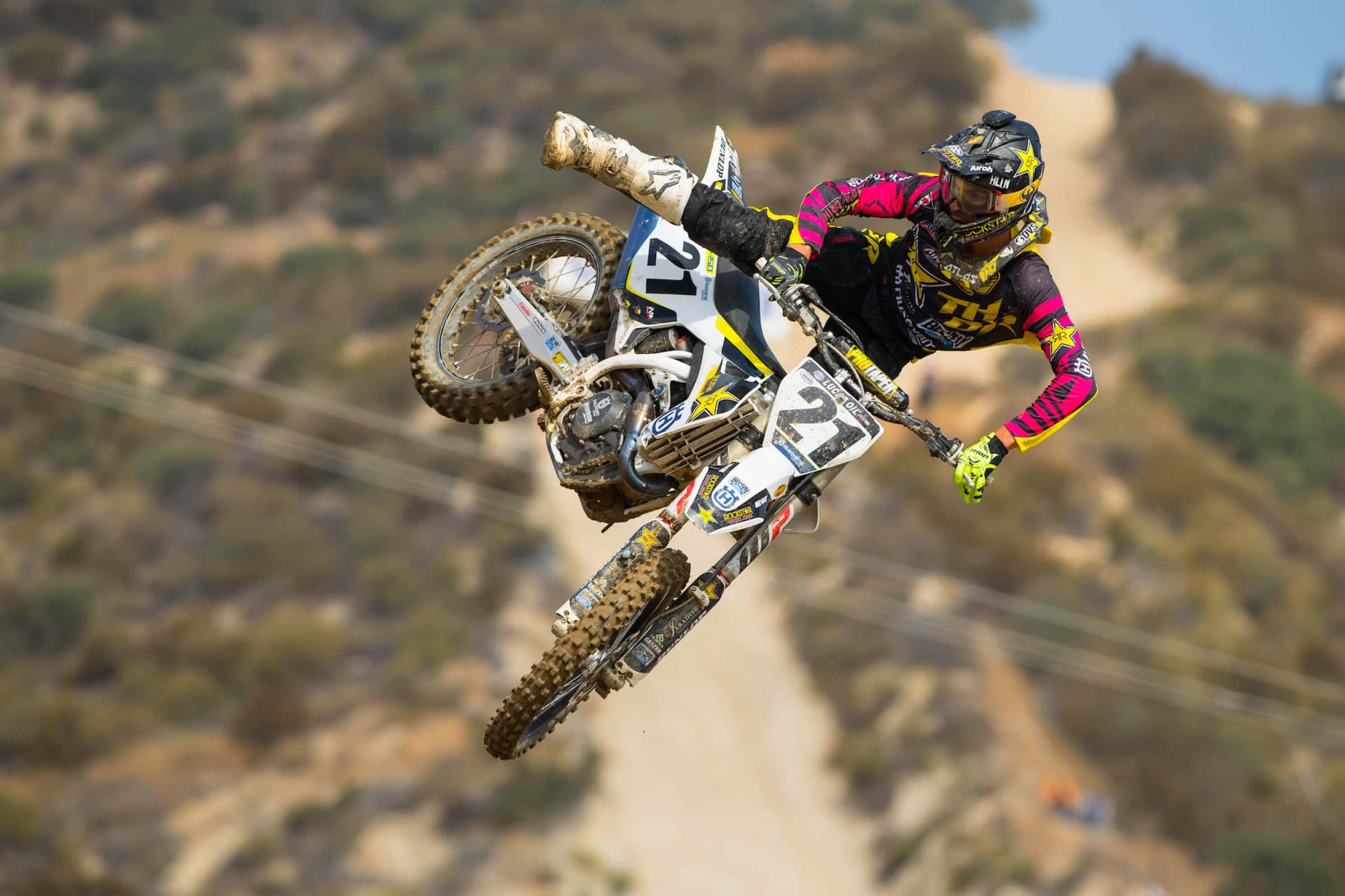 Motocross 桌布 壁紙 — 一位越野電單車手在崎嶇的泥地電單車上飛躍空中，背景是陽光照耀的山坡。