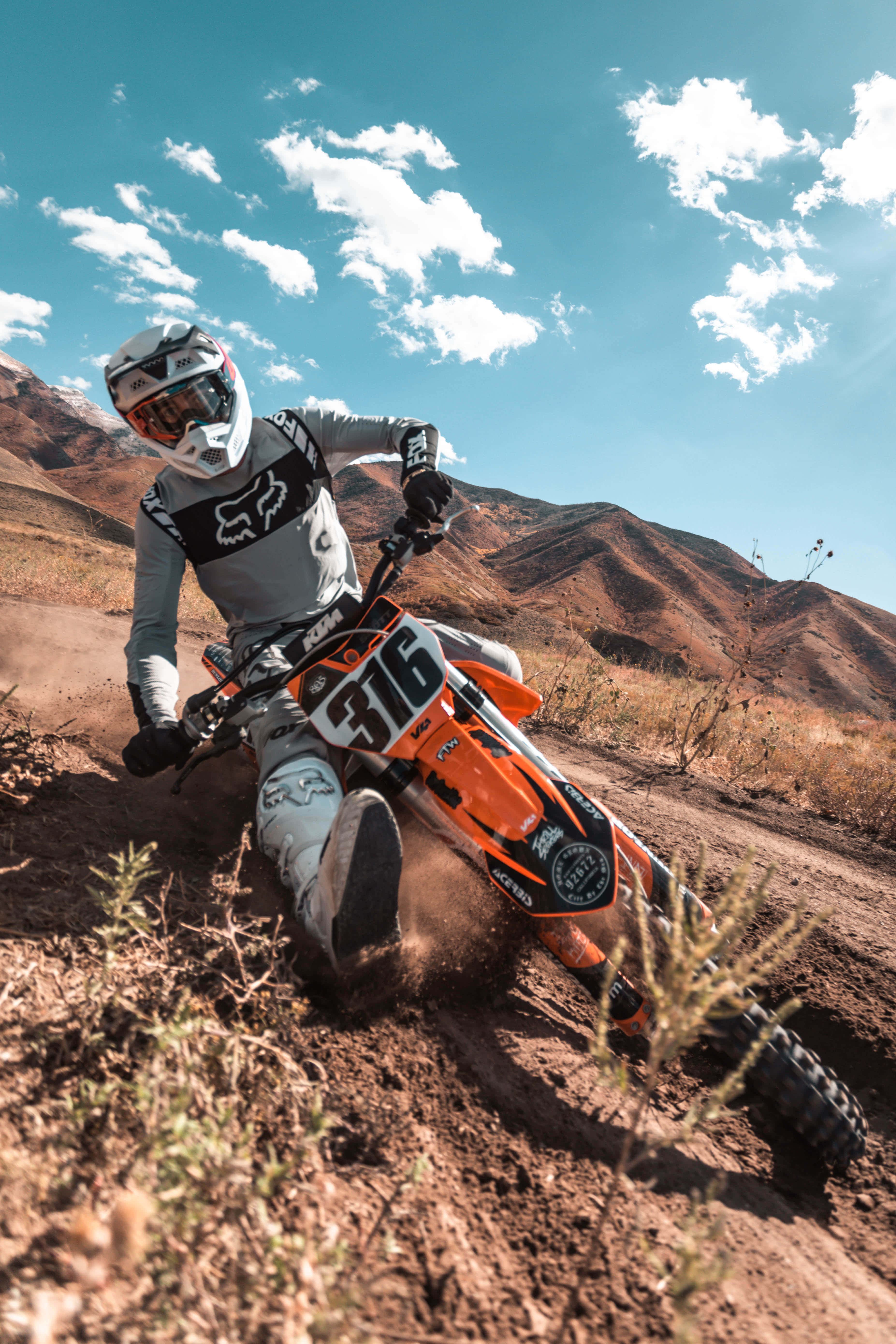 Motocross 桌布 壁紙 — 一位越野電單車手在橙色越野車上傾斜入彎，揚起塵土，背景是乾燥的山脈和藍天。