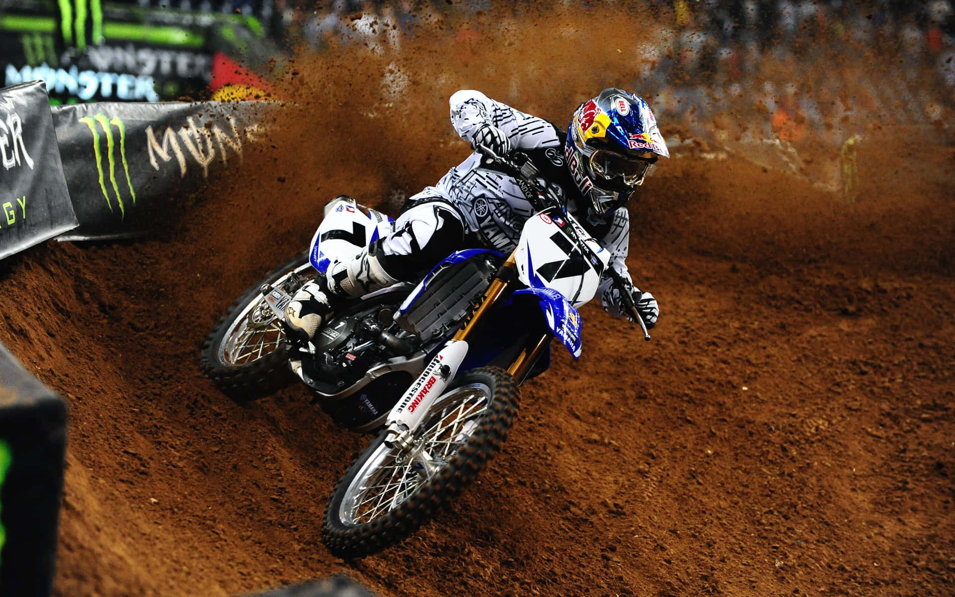Motocross 桌布 壁紙 — 一位越野電單車手在泥地賽道上傾斜入急彎，在明亮的場館燈光下揚起棕色塵土。