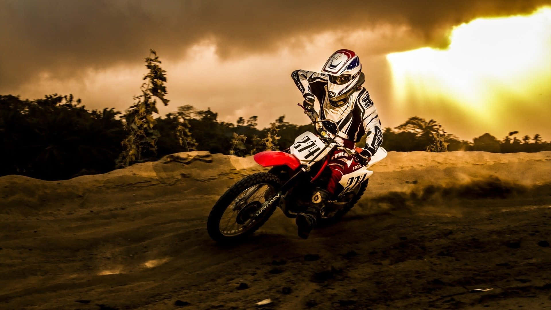 Dirtbike 桌布 壁紙 — 一位越野電單車手在泥地賽道上傾身入彎，背後的金色陽光穿過暴風雲層灑落。