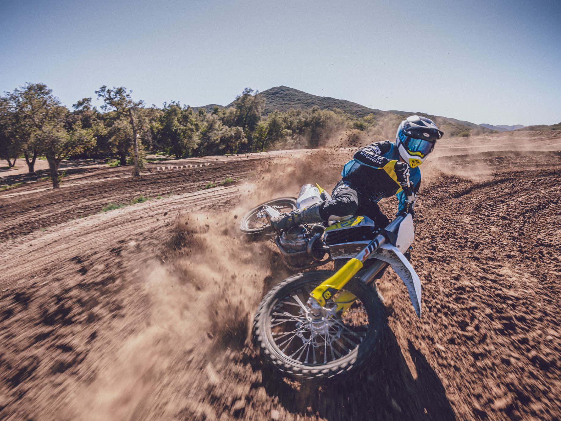 Motocross 桌布 壁紙 — 一位越野電單車手在泥地賽道上傾身過急彎，在晴朗藍天下揚起塵土。