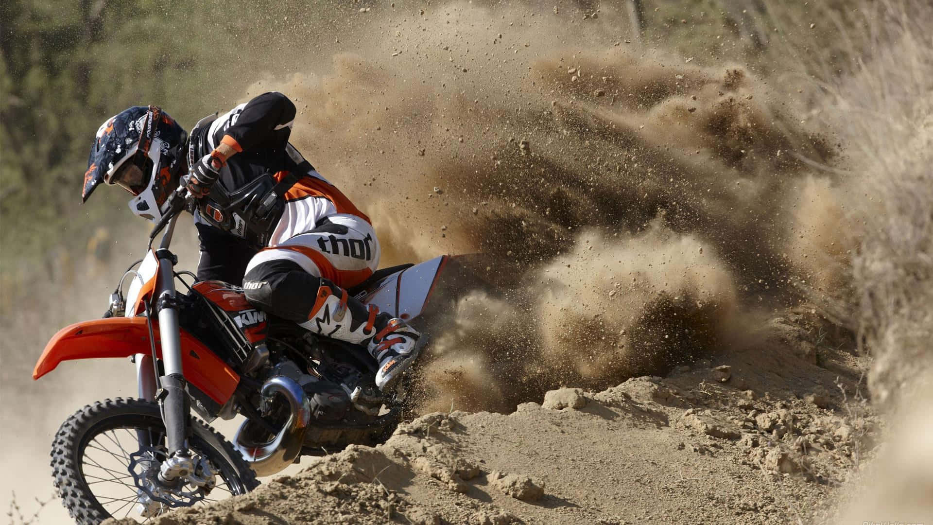 Motocross 桌布 壁紙 — 一位越野電單車手在陽光明媚的小徑上，騎著橙色越野電單車傾斜入急彎，揚起一大片棕色塵土。