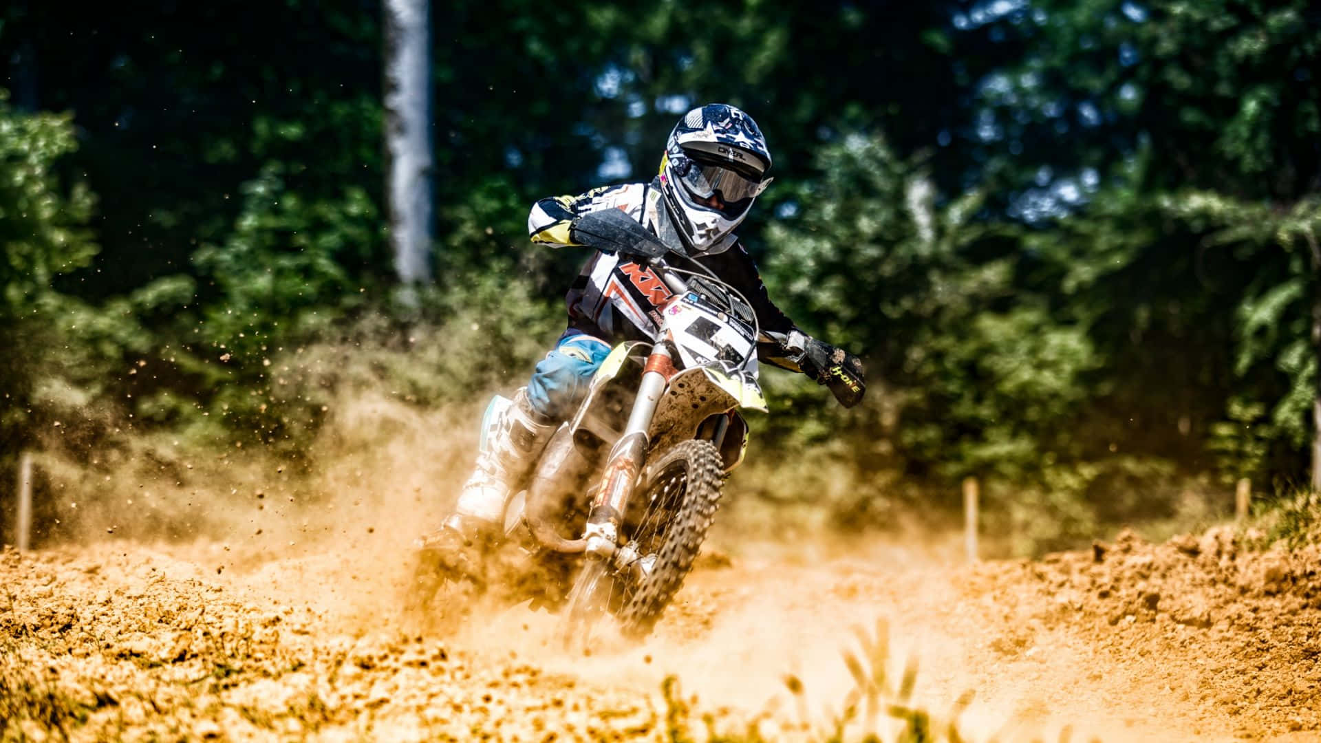 Motocross 桌布 壁紙 — 一位越野電單車手在陽光明媚的森林中的泥地賽道上傾斜入彎，揚起灰塵。