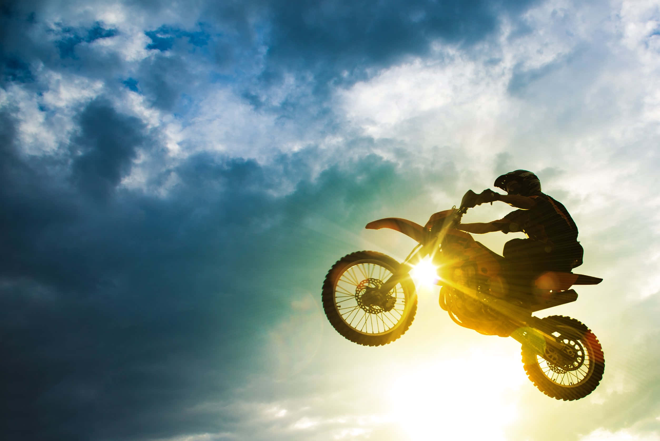 Dirtbike 桌布 壁紙 — 一位越野電單車手在雲彩和太陽眩光的戲劇性日落天空背景下翱翔於空中。