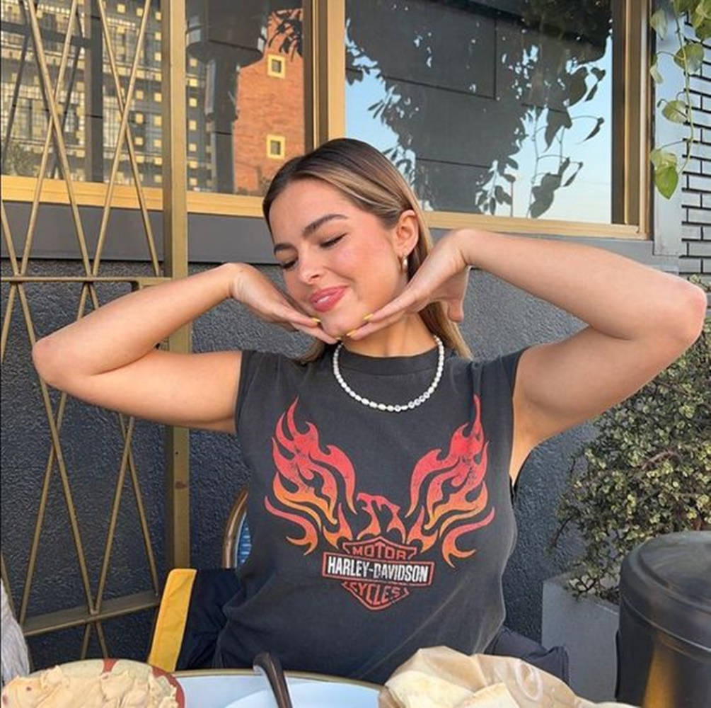 Celebrity 桌布 壁紙 — 一位身穿Harley-Davidson火焰T恤的女性在戶外咖啡館微笑，雙手框著臉部擺姿勢。