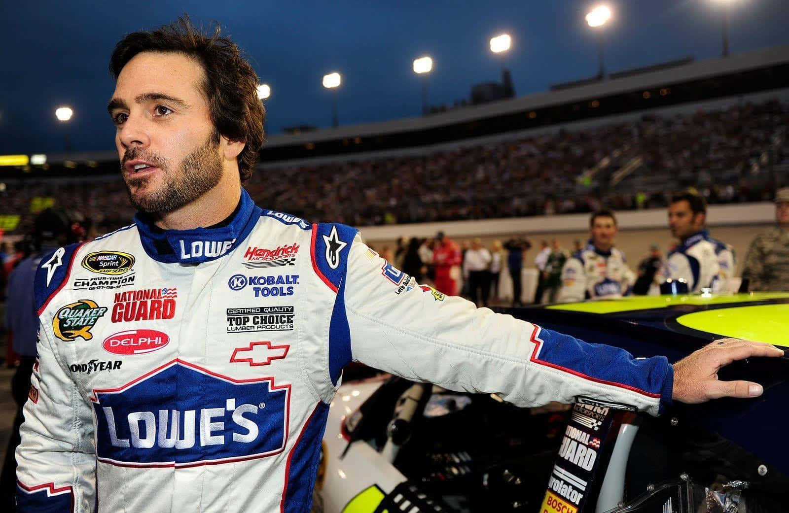 Jimmie Johnson 桌布 壁紙 — 一位身穿Lowe's賽車服的NASCAR車手在黃昏的體育場燈光下站在他的賽車旁。