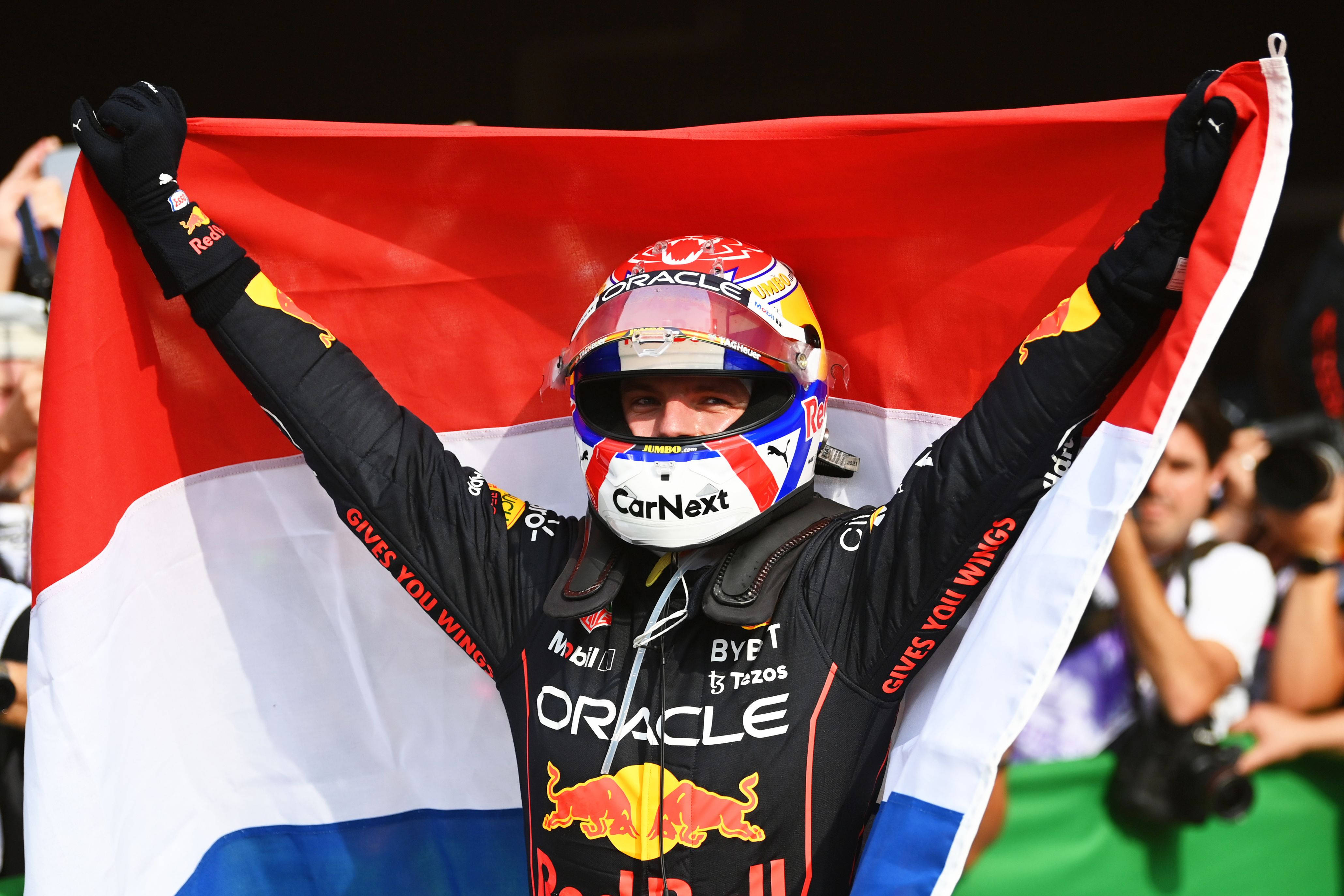 Verstappen 桌布 壁紙 — 一位身穿Red Bull賽車服和頭盔的F1車手，高舉雙臂勝利地揮舞著荷蘭國旗。