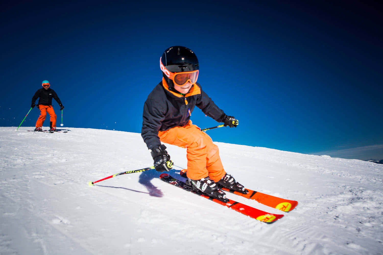 Skiing 桌布 壁紙 — 一位身穿亮橙色褲的年輕滑雪者，在晴朗藍天下沿著雪坡滑下，背景有另一位滑雪者。