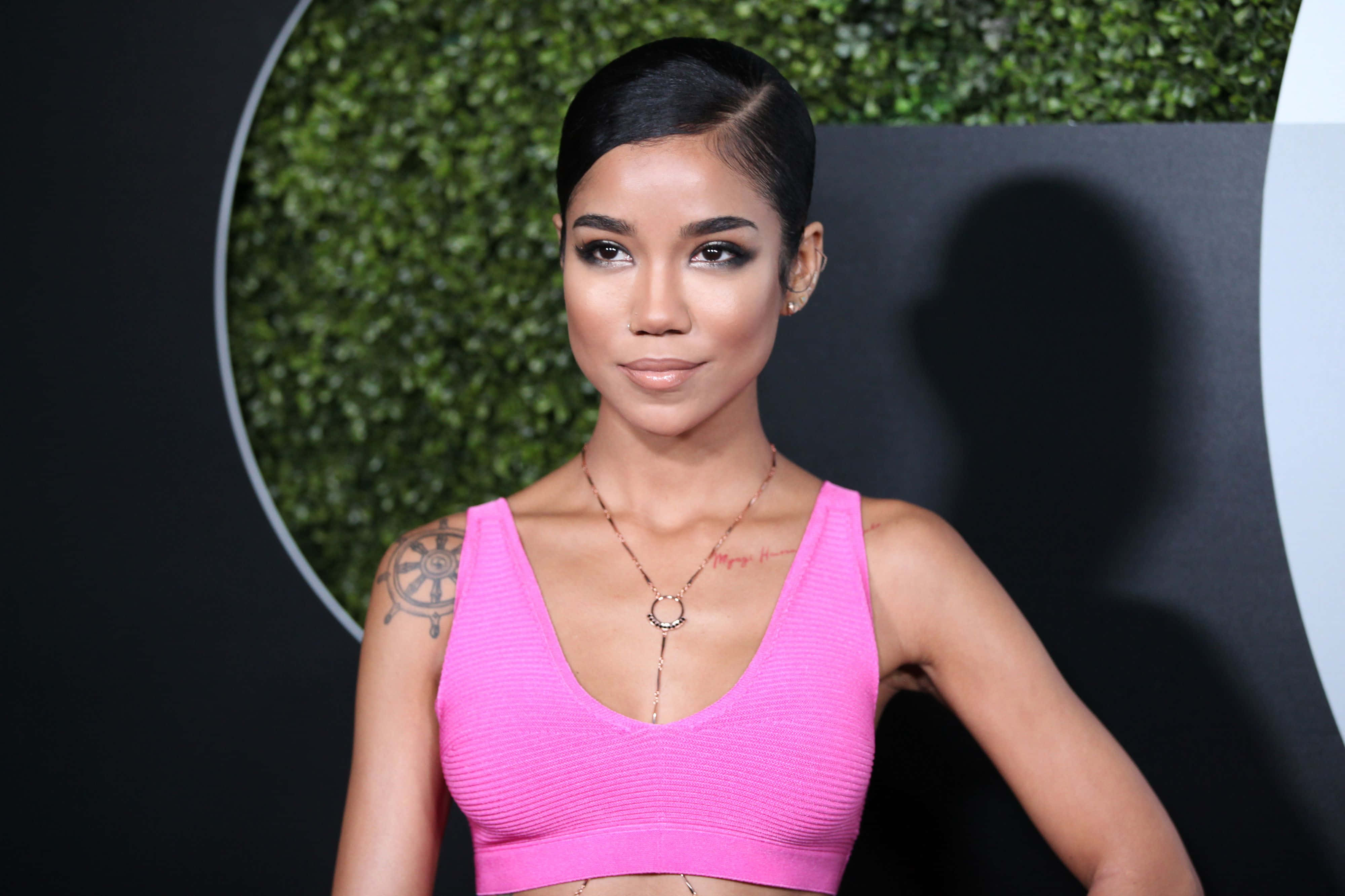 jhene aiko 桌布 壁紙 — 一位身穿亮粉紅色短版上衣的女性自信地站在黑色背景前，身後有一道圓形綠色樹籬牆。