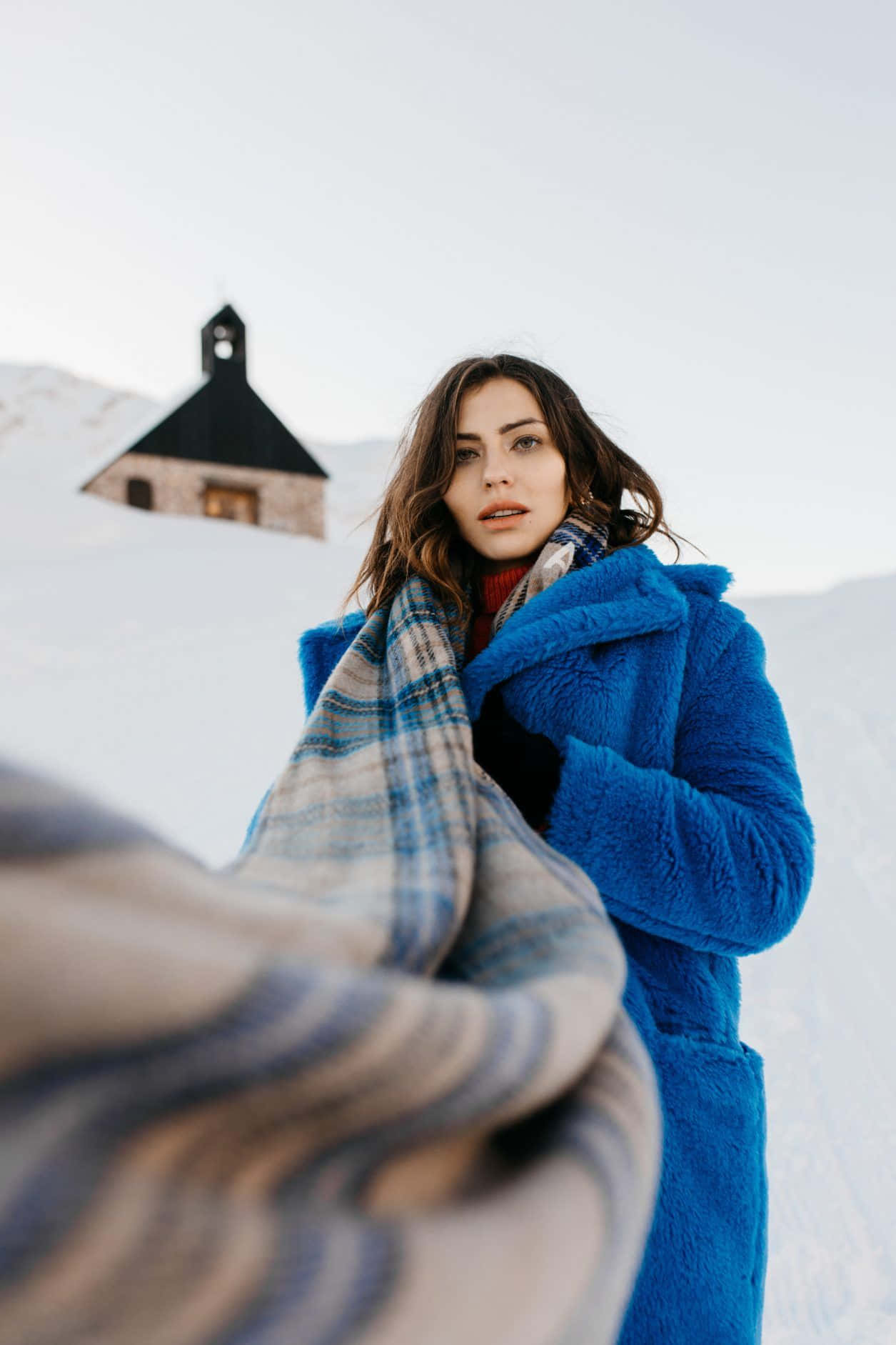 Winter Photography 桌布 壁紙 — 一位身穿亮藍色外套和格紋圍巾的女性站在雪地裡，背景是一座小教堂。