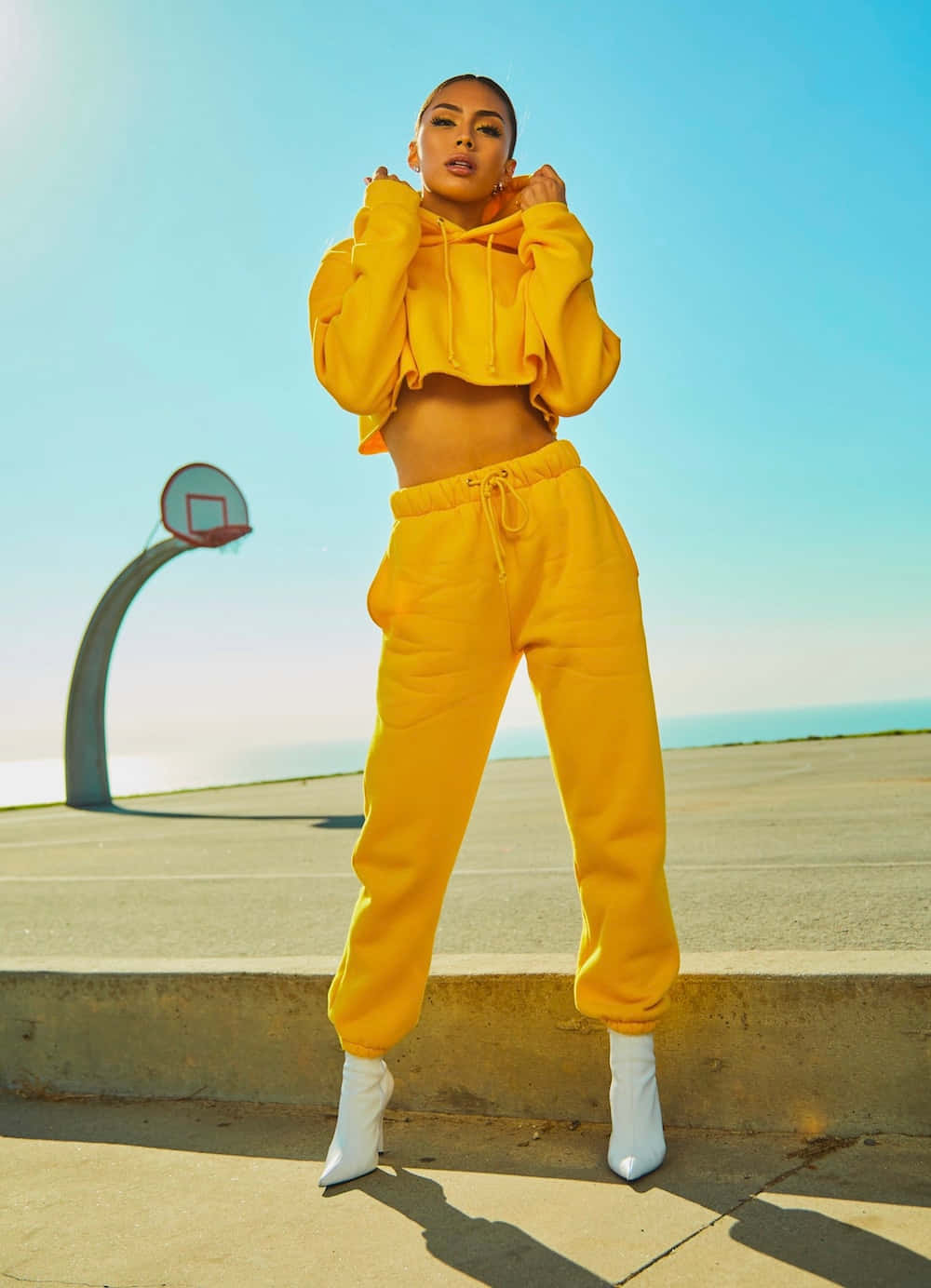 Fashion 桌布 壁紙 — 一位身穿亮黃色短版連帽衫和配套運動褲的女性，在晴朗的藍天下自信地站在籃球場上。