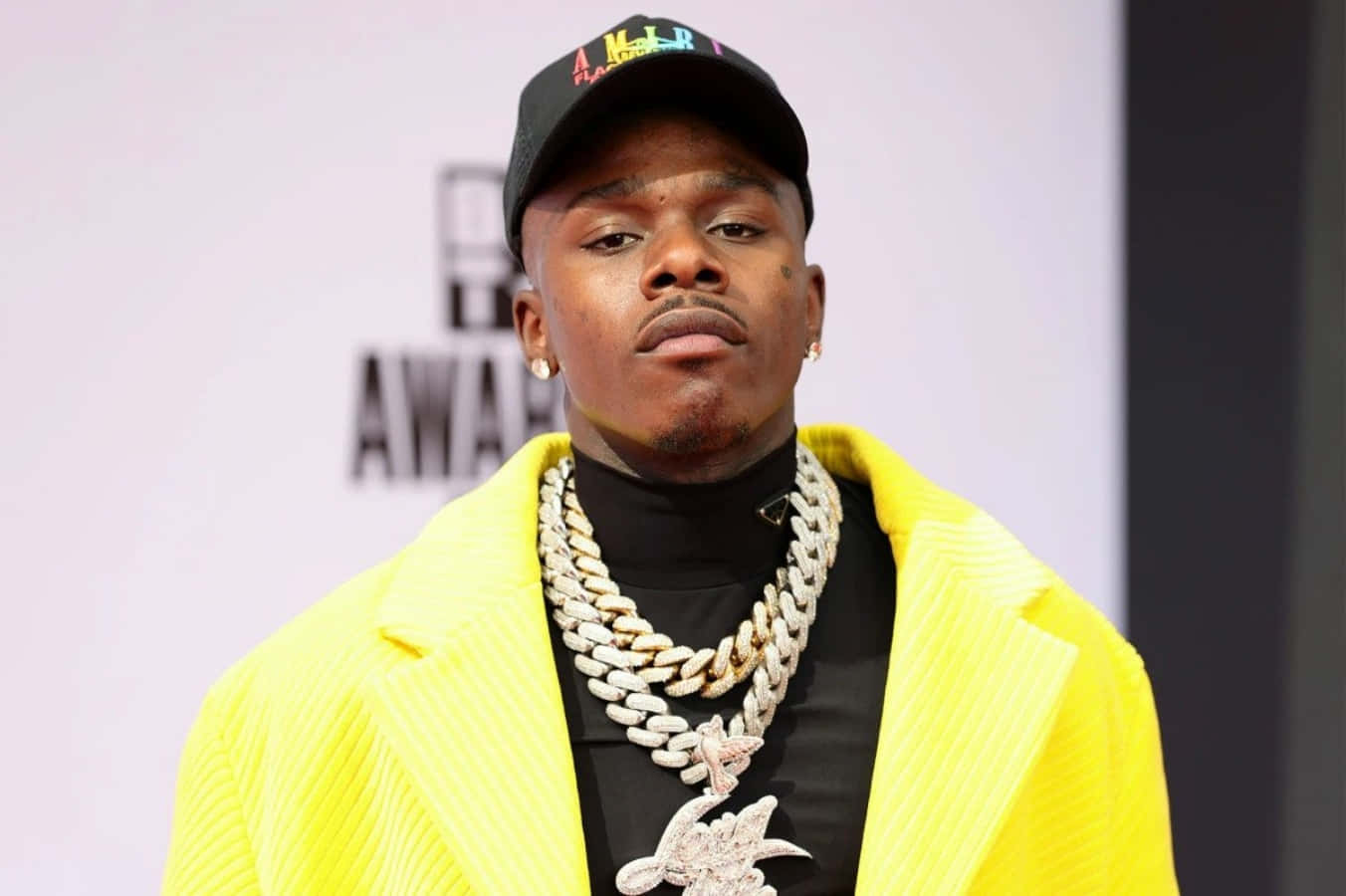 Dababy 桌布 壁紙 — 一位身穿亮黃色西裝外套和黑色帽子的男士，在頒獎典禮上佩戴大膽的鑽石鏈條自信地擺姿勢。