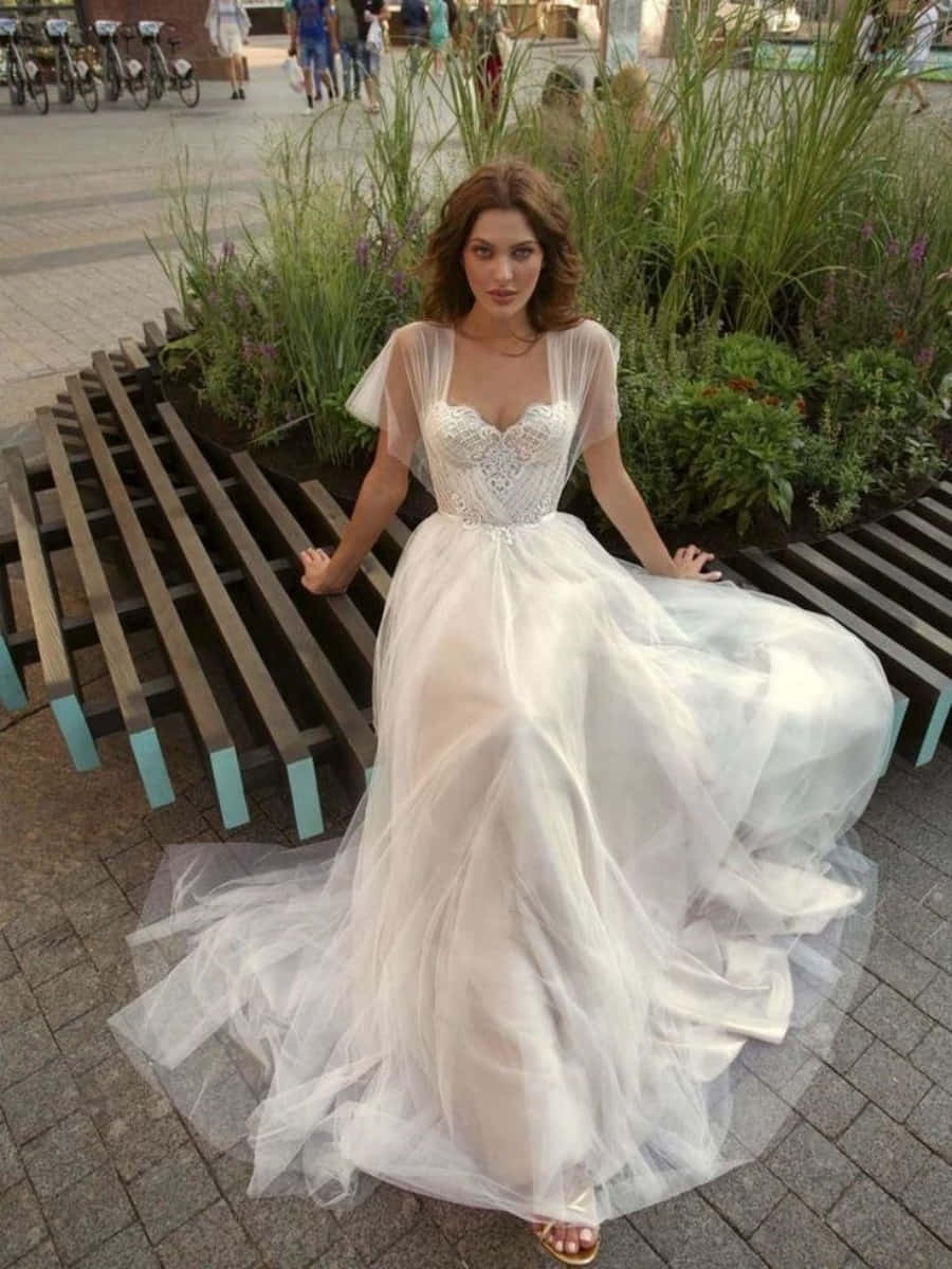 Wedding Dress 桌布 壁紙 — 一位身穿優雅白色婚紗（蕾絲上身、飄逸薄紗裙）的女性，在黃昏的城市公園中，優雅地坐在木長椅上擺姿勢。