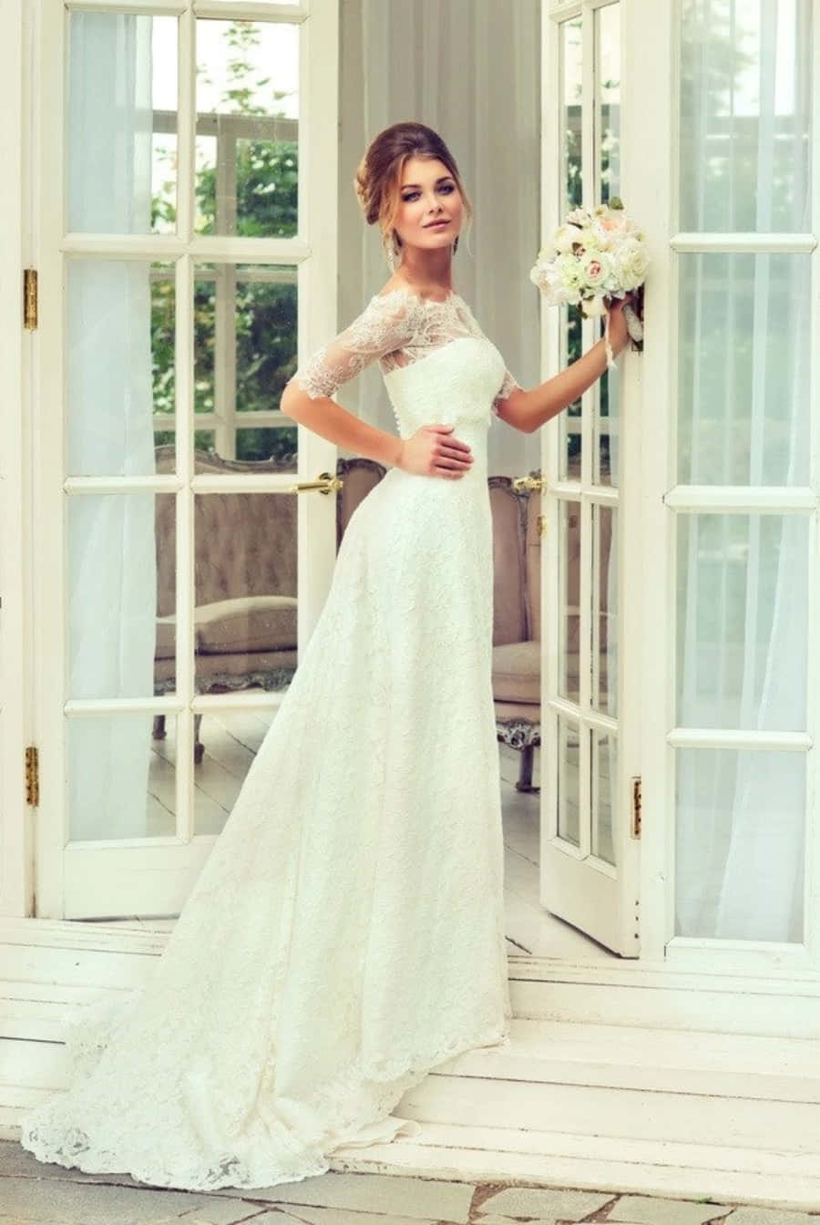 Wedding Dress 桌布 壁紙 — 一位身穿優雅白色蕾絲禮服的新娘，優雅地站在門廊上，手持花束，靠著法式門。