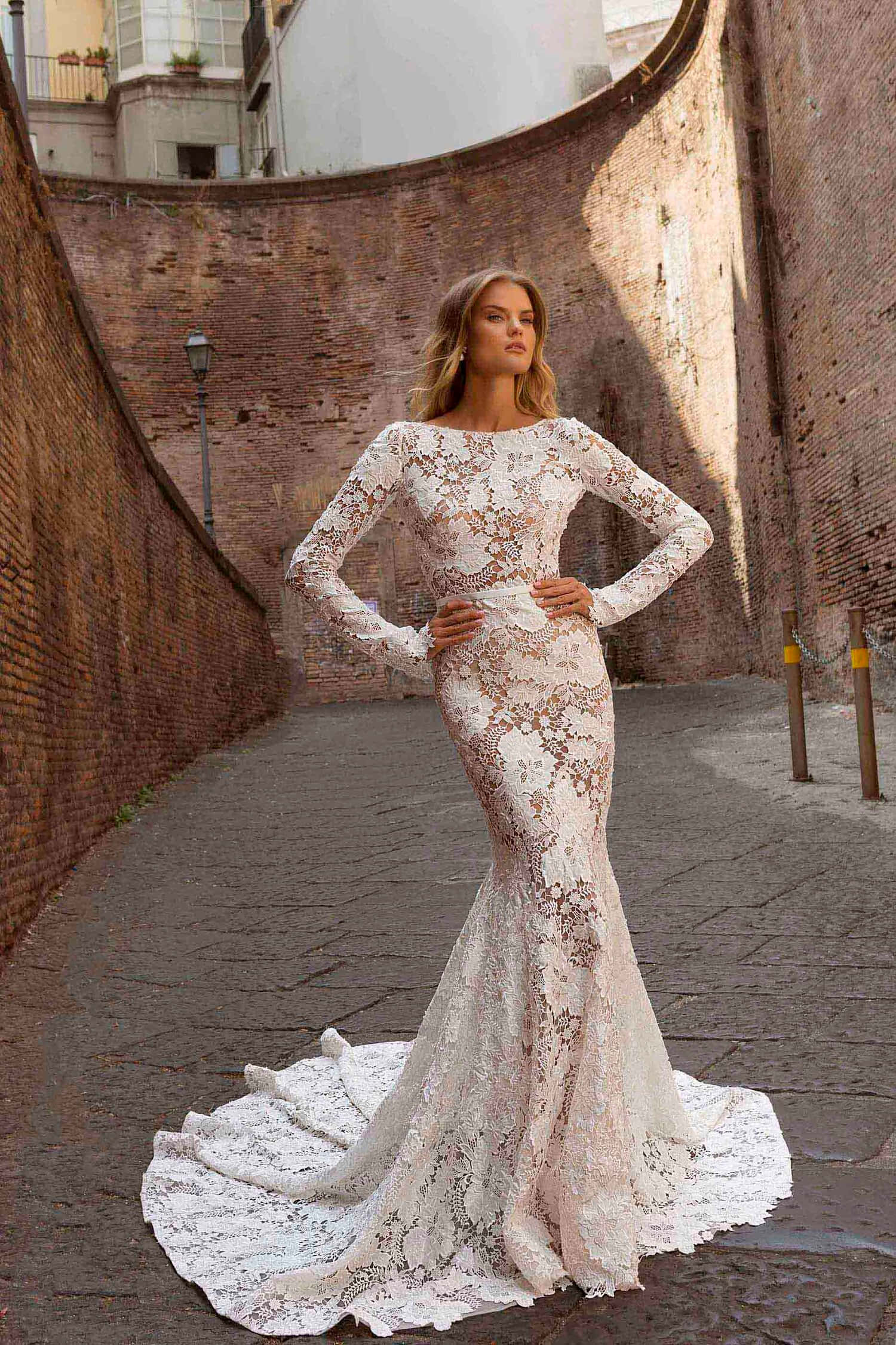 Bride's Dress 桌布 壁紙 — 一位身穿優雅白色蕾絲美人魚禮服的女士自信地站在陽光普照的狹窄磚砌小巷中。