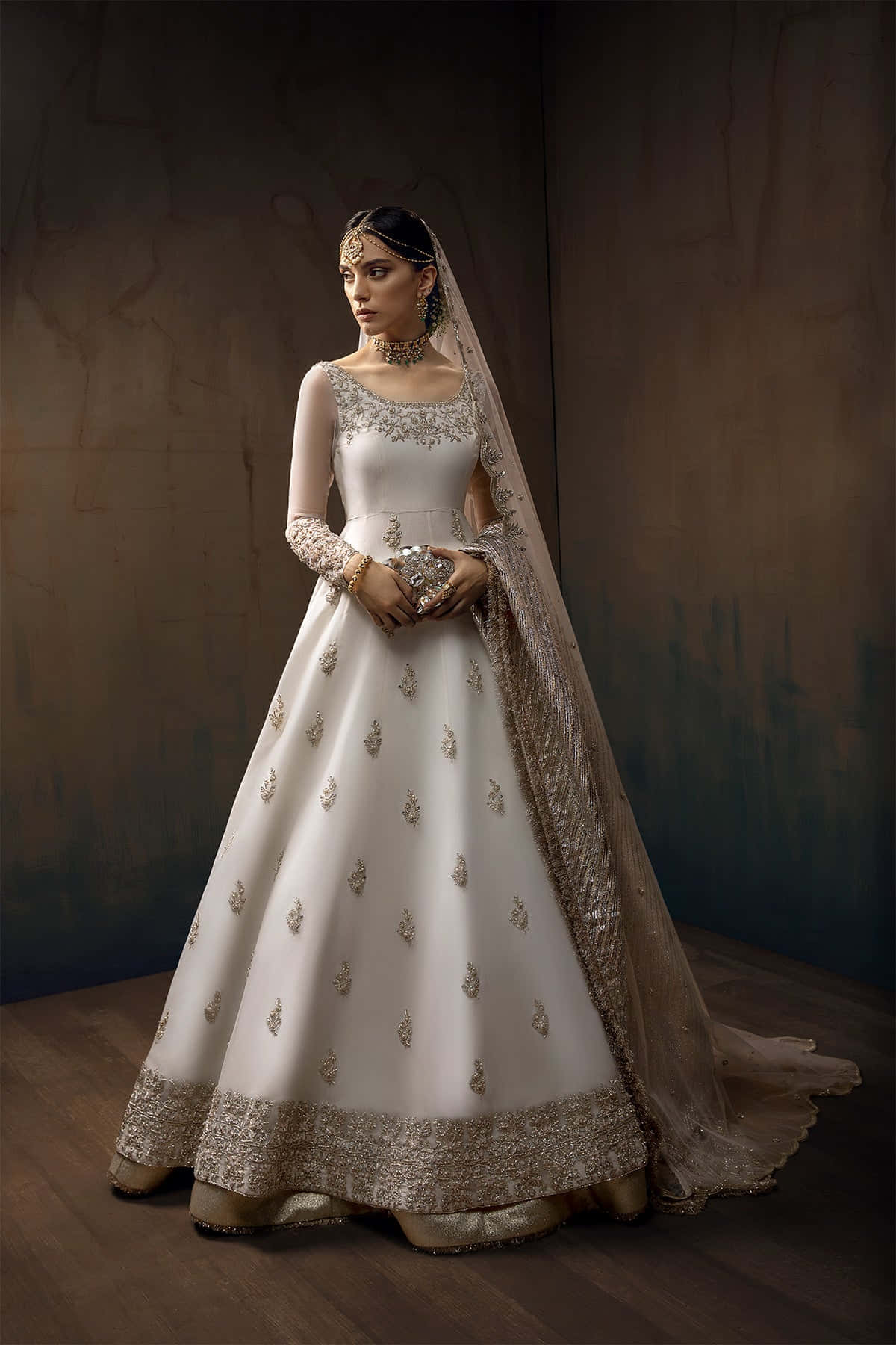 Bride's Dress 桌布 壁紙 — 一位身穿優雅象牙色刺繡長裙、飾有金色細節的女性，在質感深色牆壁前從容站立。
