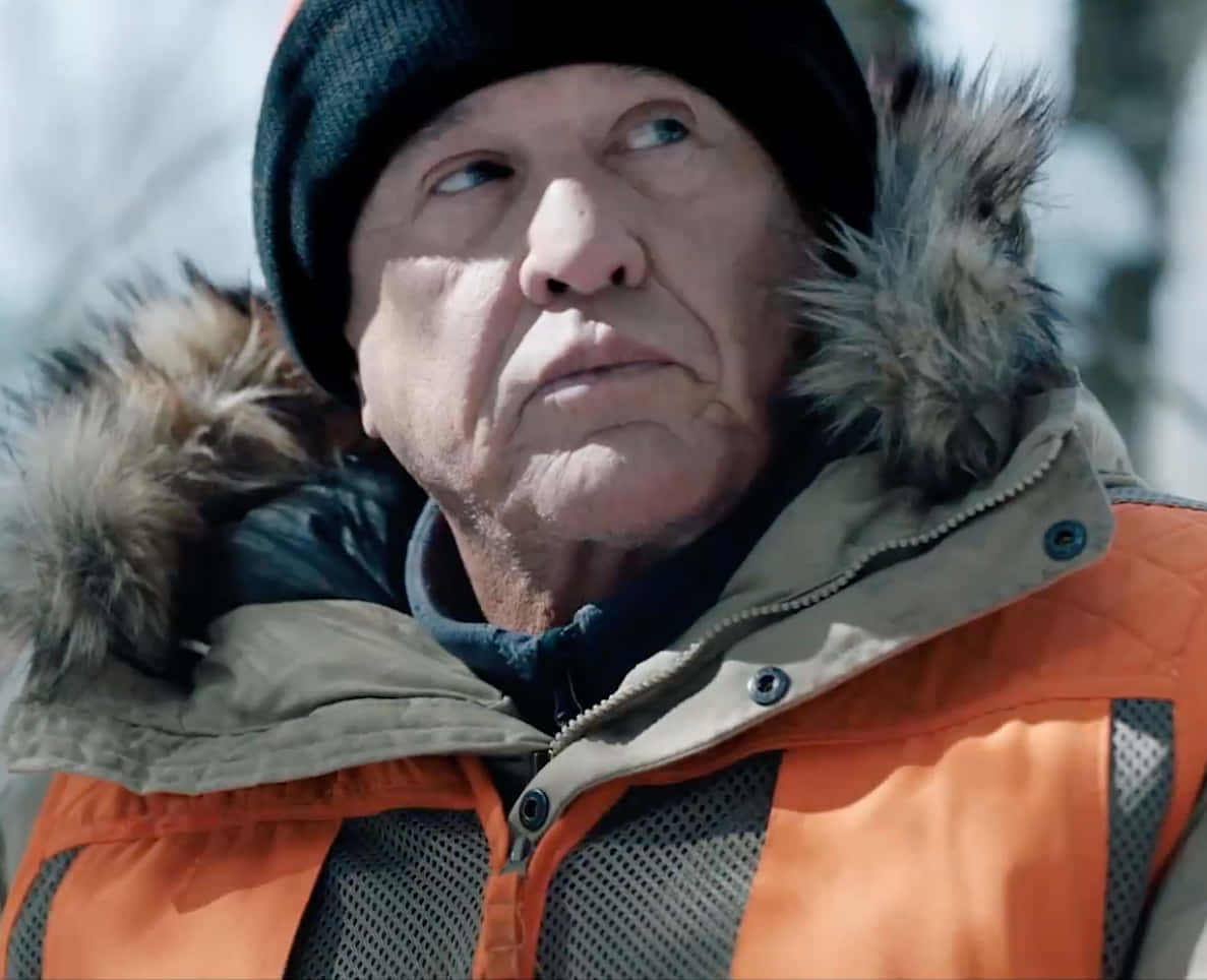 Tom Berenger 桌布 壁紙 — 一位身穿冬季外套和冷帽的長者，在雪景背景下帶著嚴肅的表情向上凝視。