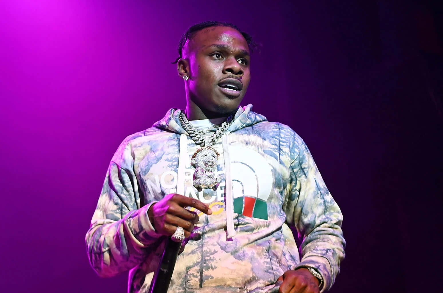 Dababy 桌布 壁紙 — 一位身穿圖案連帽衫和鏈條的男性表演者站在充滿活力的紫色舞台燈光下，手持麥克風。