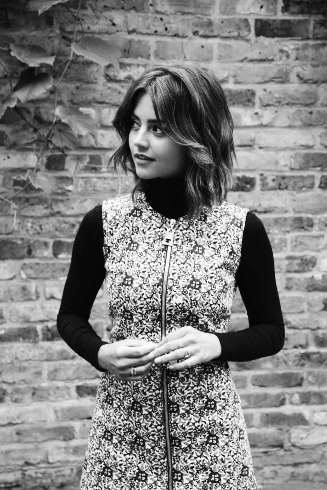 Jenna Coleman 桌布 壁紙 — 一位身穿圖案連衣裙的女性站在佈滿常春藤的風化磚牆前，以引人注目的黑白影像捕捉。