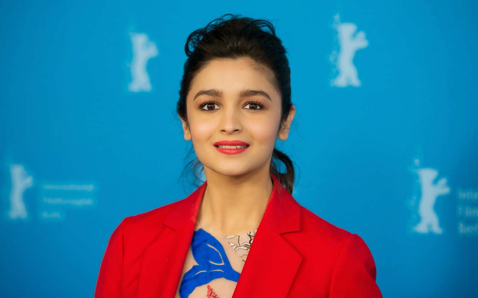 Alia Bhatt 桌布 壁紙 — 一位身穿大膽紅色西裝外套的女性，在帶有白色熊剪影的鮮豔藍色背景前溫暖地微笑。