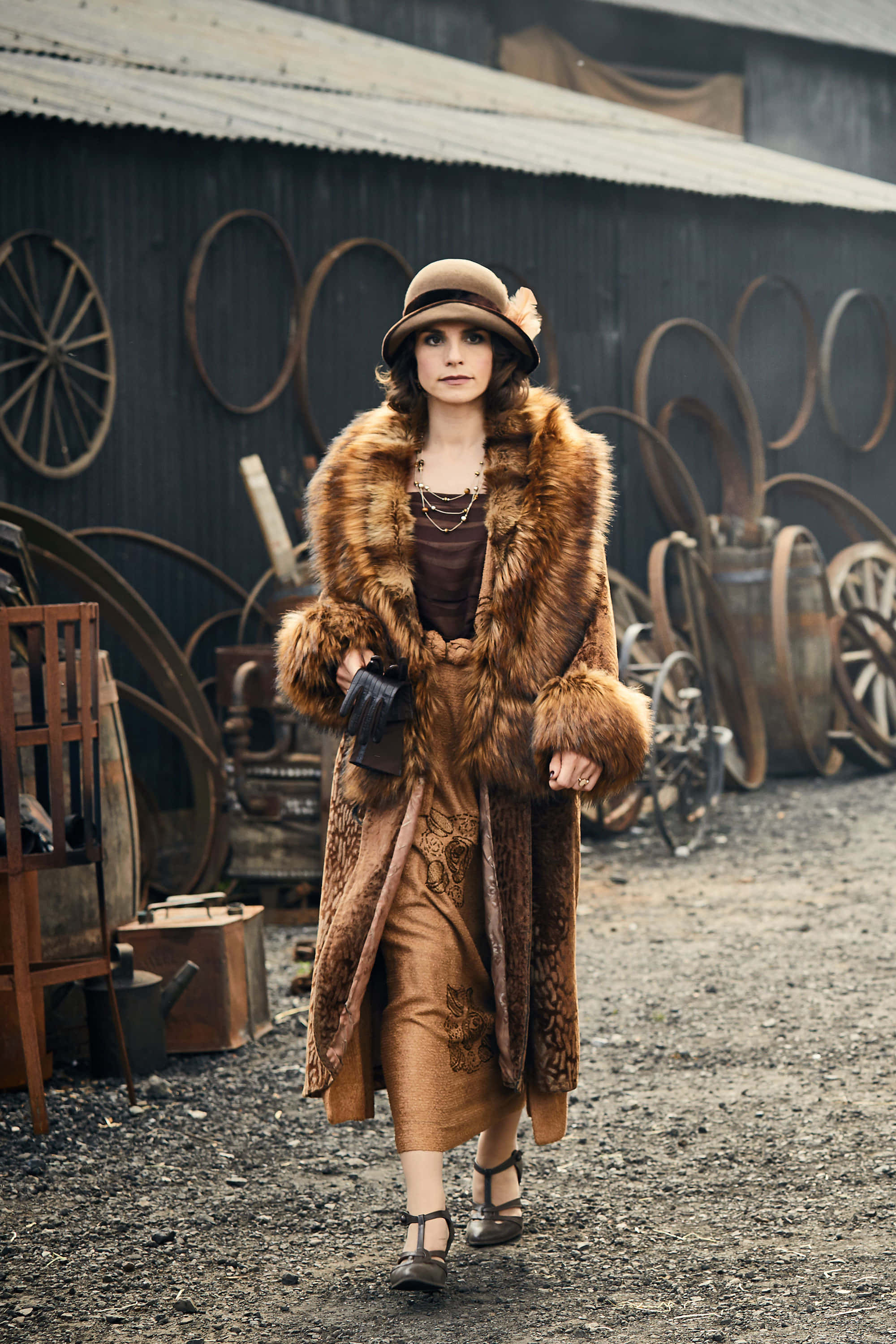 Peaky Blinders 桌布 壁紙 — 一位身穿奢華棕色皮草大衣和鐘形帽的女性自信地走過一個骯髒工業場地中的鄉村農場設備。
