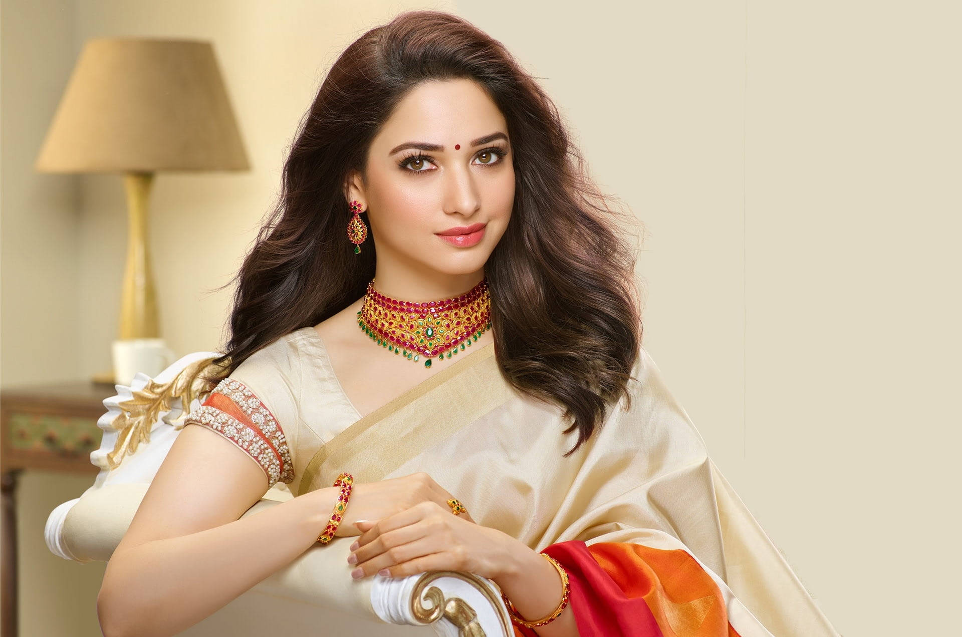 Bollywood 桌布 壁紙 — 一位身穿奶油色絲綢紗麗、佩戴紅色和金色珠寶的女性，在柔和燈光的房間裡優雅地擺姿勢在燈旁。