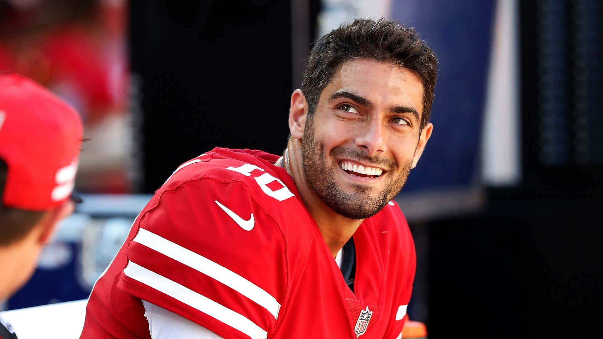 Jimmy Garoppolo 桌布 壁紙 — 一位身穿帶有白色條紋的紅色球衣、面帶微笑的足球運動員，帶著明亮的表情看向一旁。