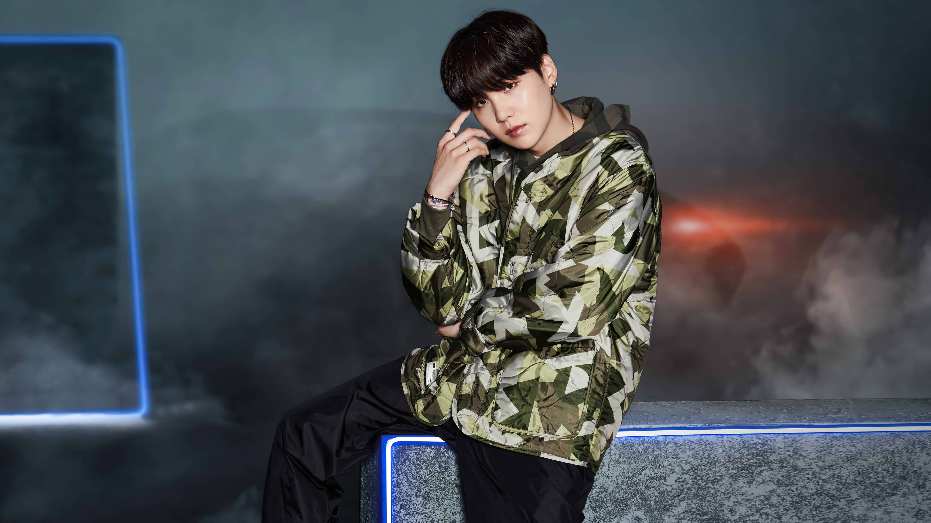 BTS SUGA For Fila 壁紙 — 一位身穿幾何迷彩外套的年輕男子自信地坐在發光的藍色邊緣上，背景是煙霧瀰漫的黑暗環境和霓虹燈。