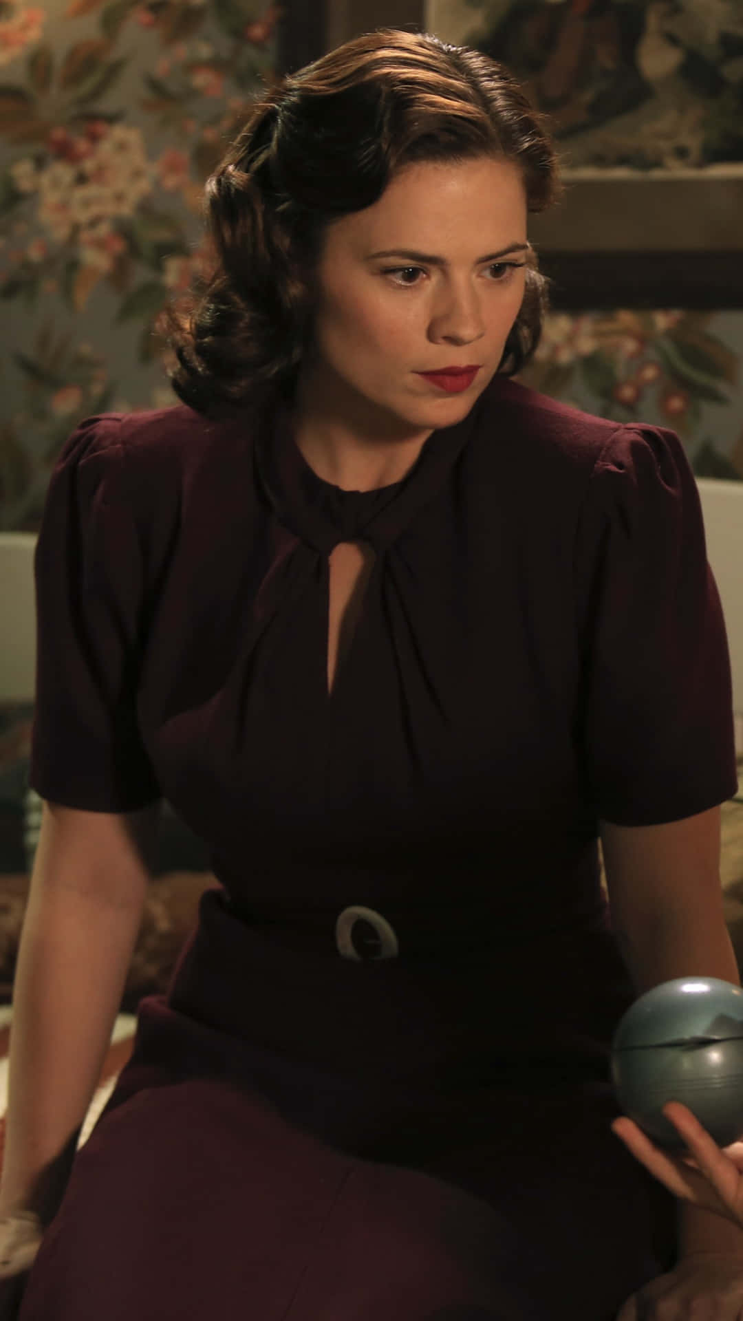 Peggy Carter 桌布 壁紙 — 一位身穿復古酒紅色連衣裙、領口有鑰匙孔設計的女性，坐在花卉牆紙背景前，手持一個金屬球體。