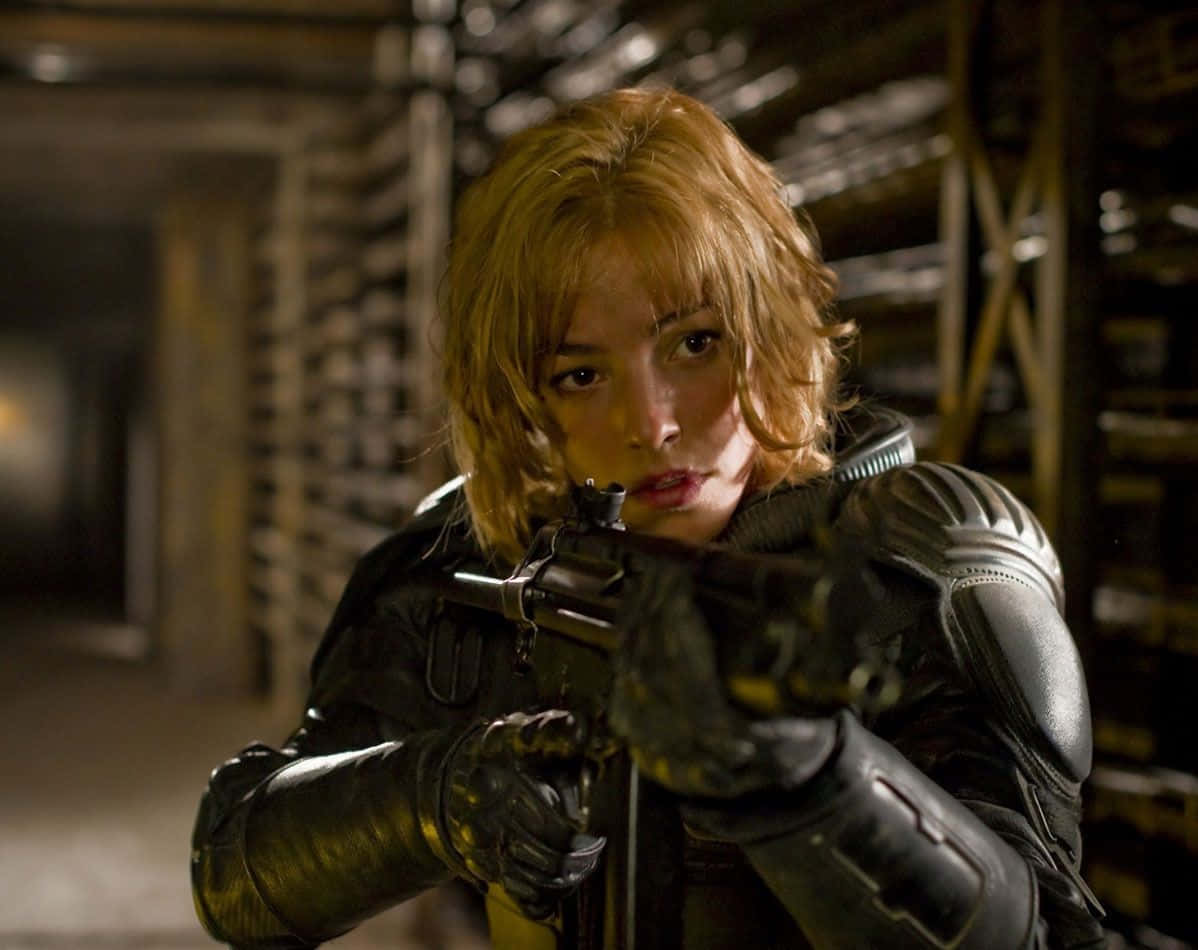 Olivia Thirlby 桌布 壁紙 — 一位身穿戰術裝備的堅定女性，在光線昏暗的工業倉庫中，全神貫注地瞄準步槍。
