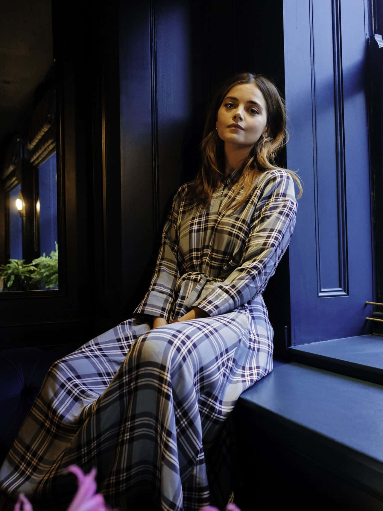 Jenna Coleman 桌布 壁紙 — 一位身穿格仔裙嘅女人沉思咁坐喺藍色窗台上，沐浴喺柔和嘅晚光中。