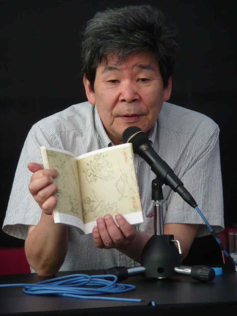 Isao Takahata 桌布 壁紙 — 一位身穿條紋襯衫的年長男性在深色背景前，手持打開的素描本對著麥克風。
