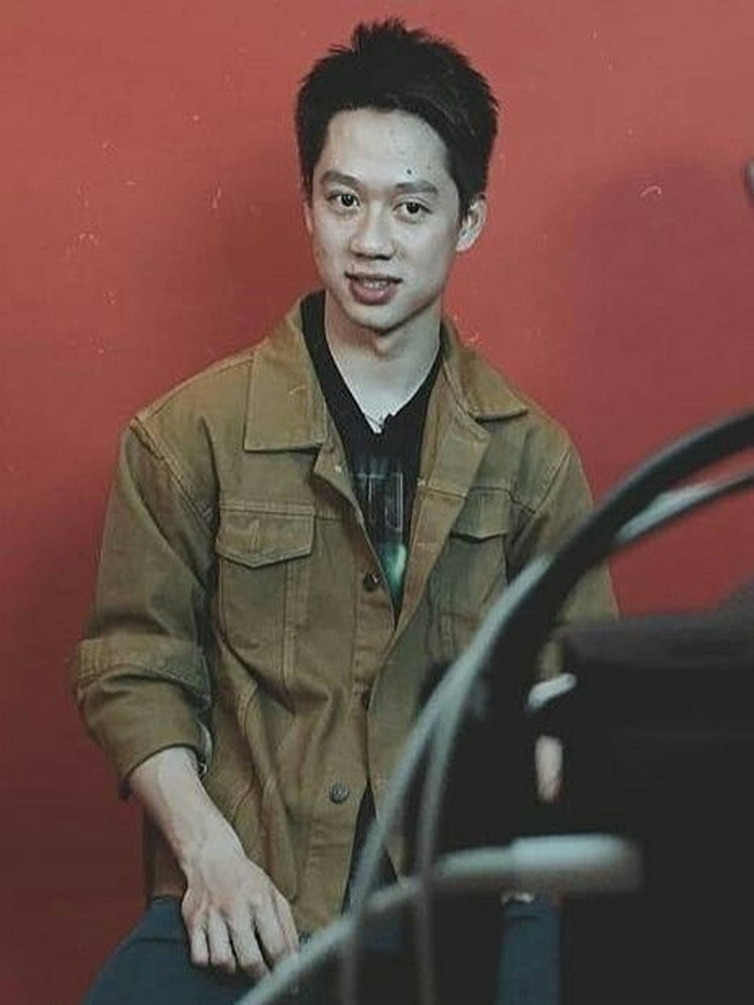 桌布 壁紙 — 一位身穿棕色外套的年輕男子，在帶有紋理的紅色牆壁前微笑，前景元素模糊。