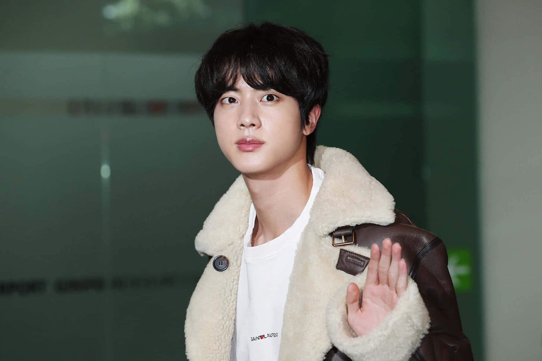 Jin 桌布 壁紙 — 一位身穿棕色羊羔絨外套的年輕男子站在室內玻璃門附近揮手。