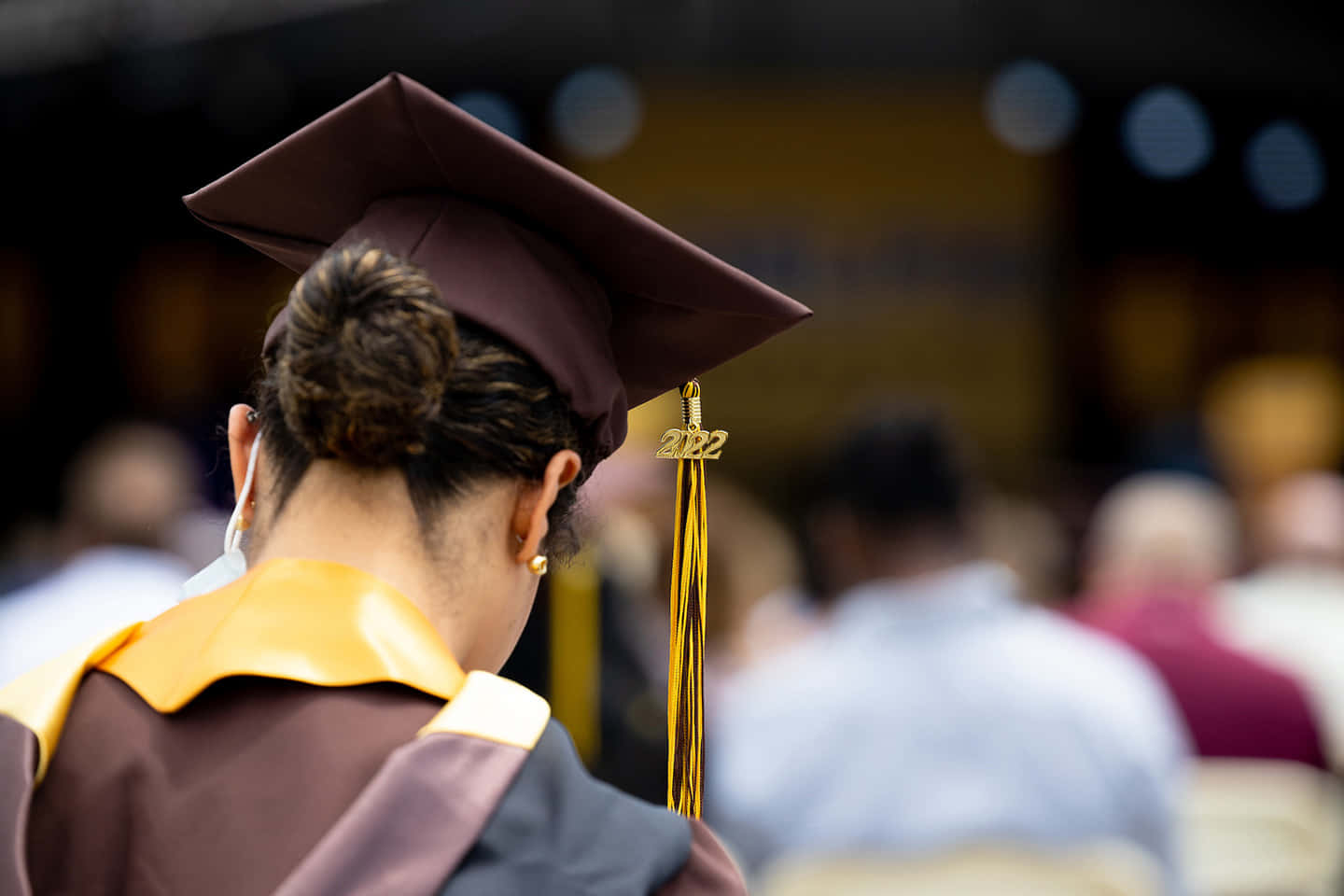 Graduation Ceremony 桌布 壁紙 — 一位身穿棗紅色畢業袍、頭戴2022金色流蘇四方帽的畢業生，低頭看著背景模糊的典禮人群。