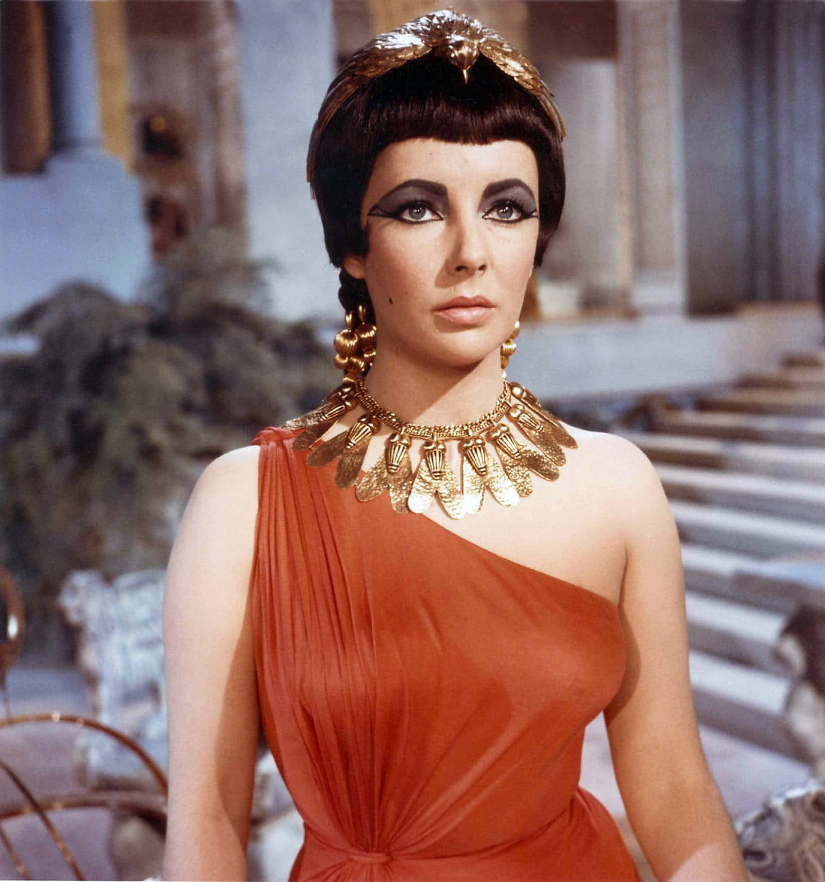 Cleopatra 桌布 壁紙 — 一位身穿橙色單肩長裙和金色珠寶的女性，威嚴地站在古典石庭院中。