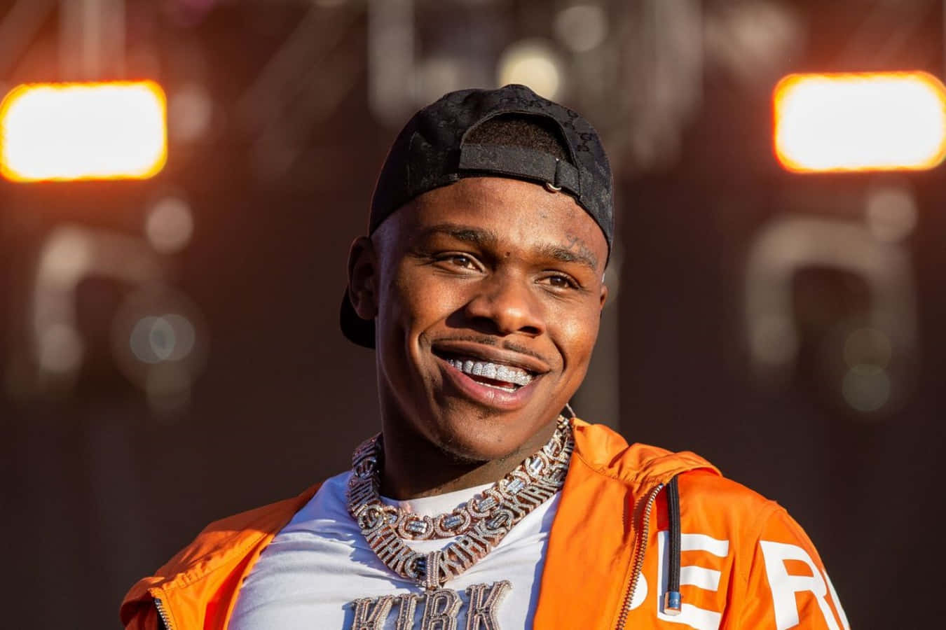 Dababy 桌布 壁紙 — 一位身穿橙色外套和黑色帽子的微笑男子，在溫暖的舞台燈光下對著鏡頭綻放笑容。