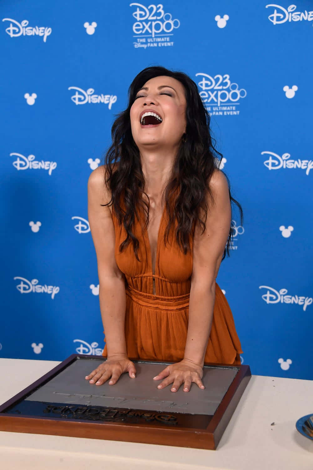 Ming Na Wen 桌布 壁紙 — 一位身穿橙色連衣裙的女性在D23 Expo活動中，將雙手按在迪士尼手印牌上，開懷大笑。