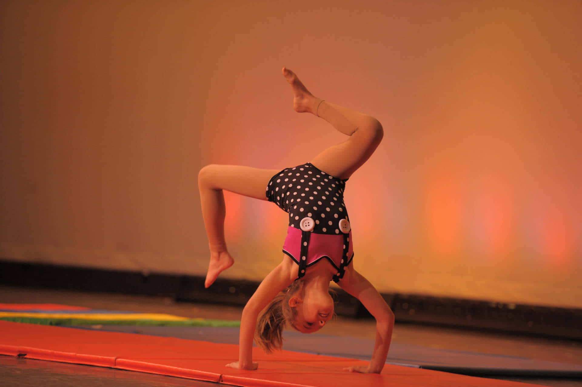 Gymnastics 桌布 壁紙 — 一位身穿波點緊身衣的小女孩在色彩繽紛的體操墊上，於溫暖的舞台燈光下做出充滿動感的手倒立。