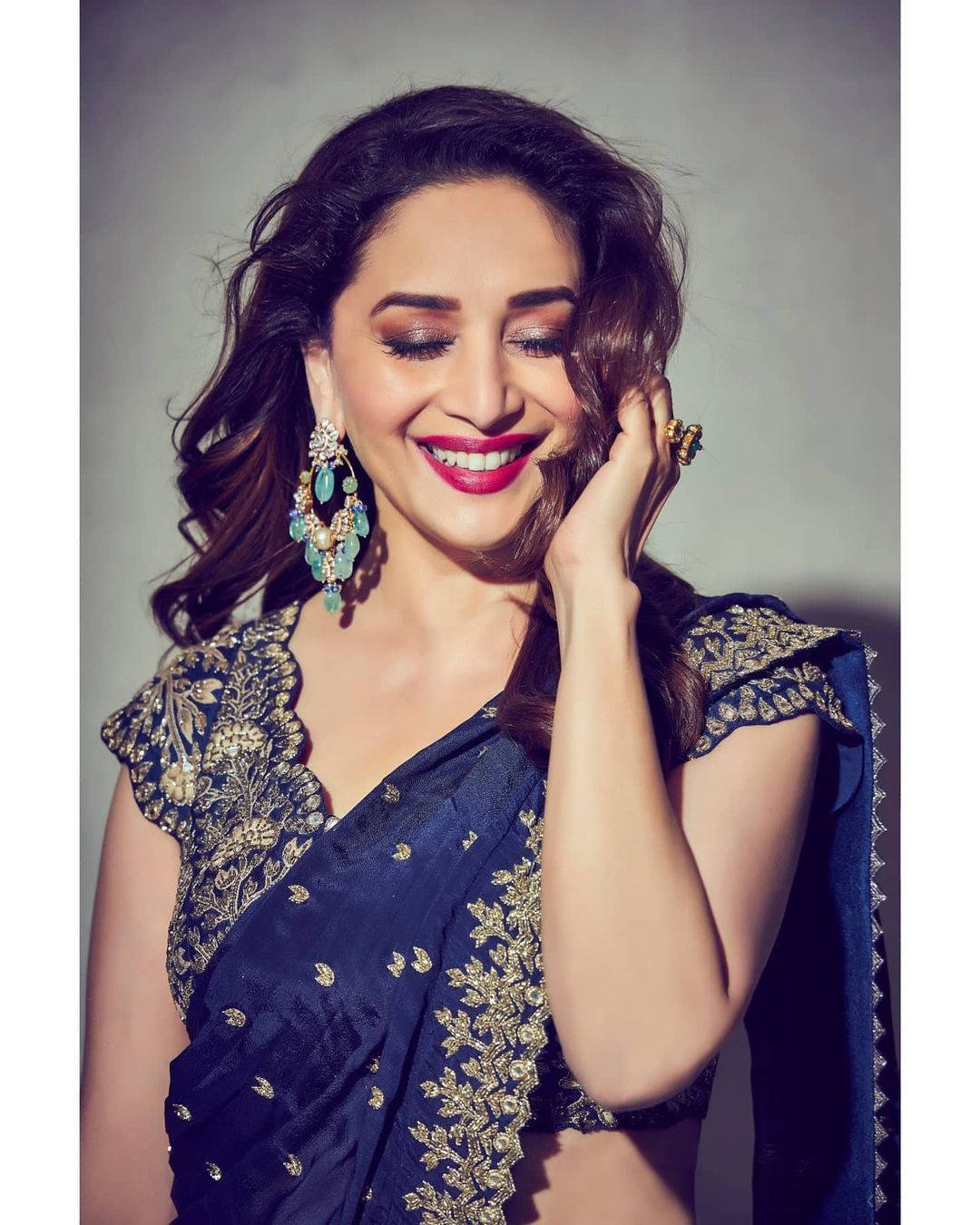 Madhuri Dixit 桌布 壁紙 — 一位身穿海軍藍刺繡紗麗的容光煥發的女性，一邊用手觸摸臉頰，一邊溫暖地微笑。