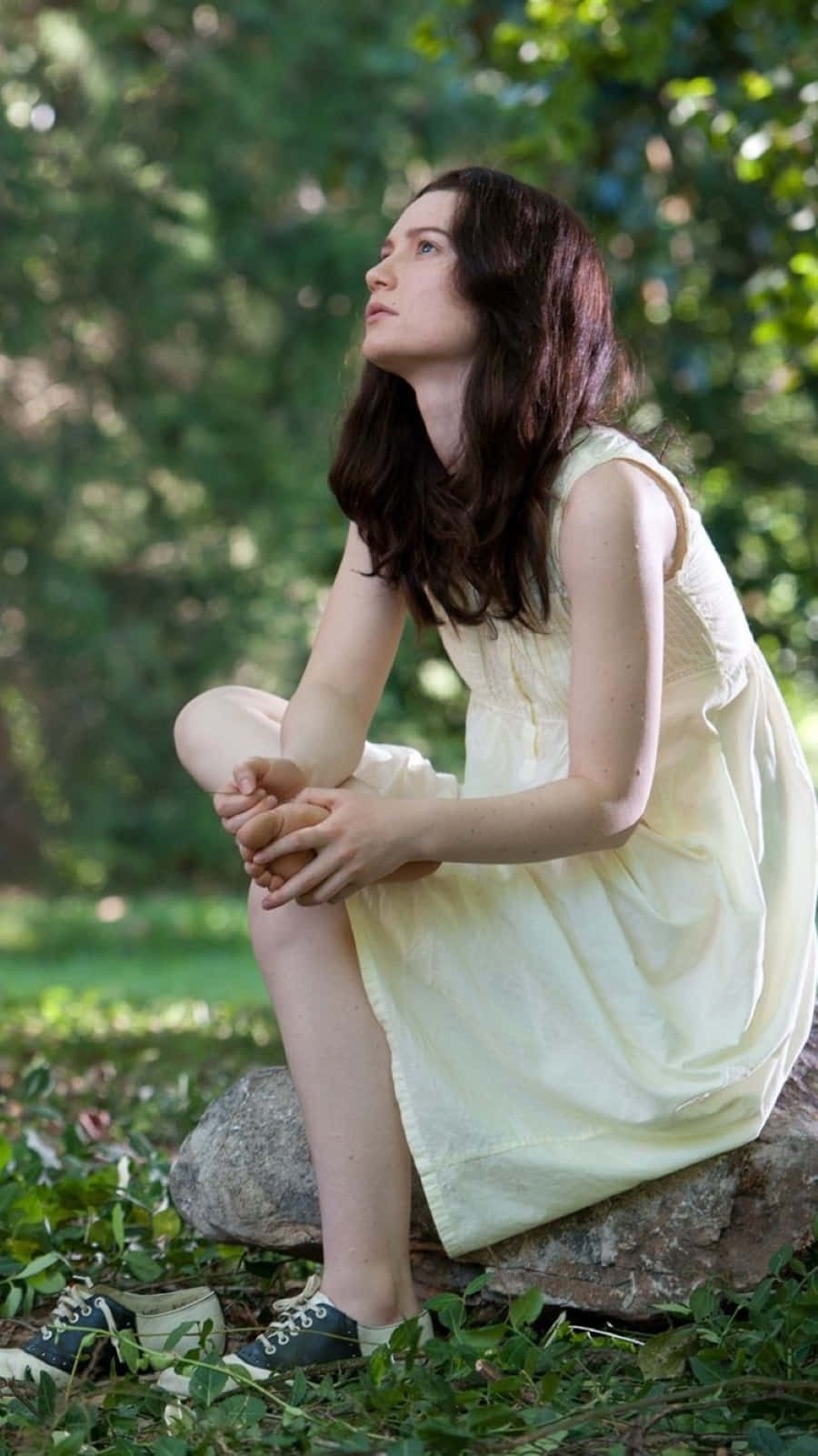 Mia Wasikowska 桌布 壁紙 — 一位身穿淡黃色連衣裙的年輕女子坐在長滿苔蘚的石頭上，周圍是茂密的綠色樹葉，若有所思。
