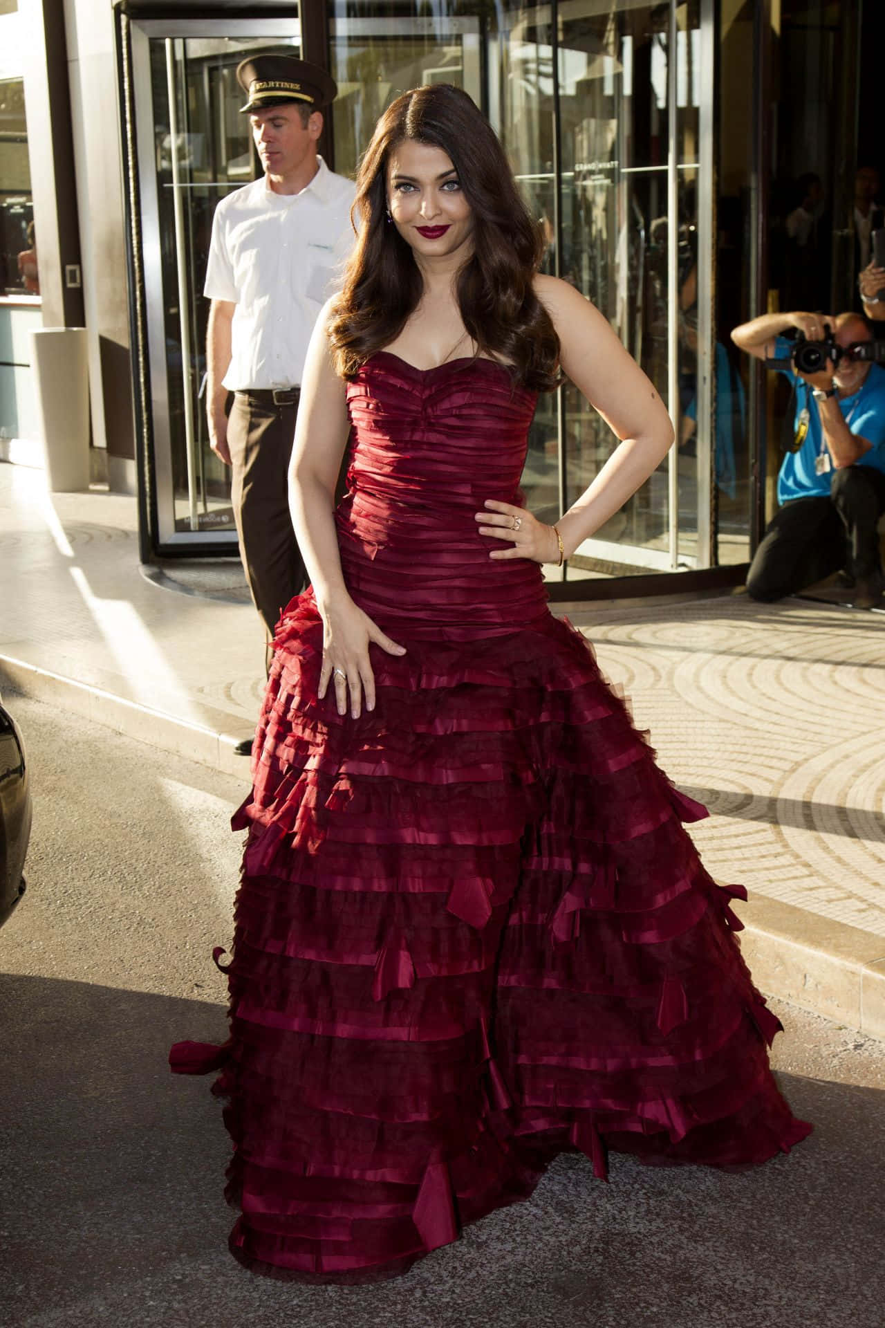 Aishwarya Rai 桌布 壁紙 — 一位身穿深紅色荷葉邊連衣裙的女性自信地在現代玻璃建築外擺姿勢，附近有攝影師和門衛。