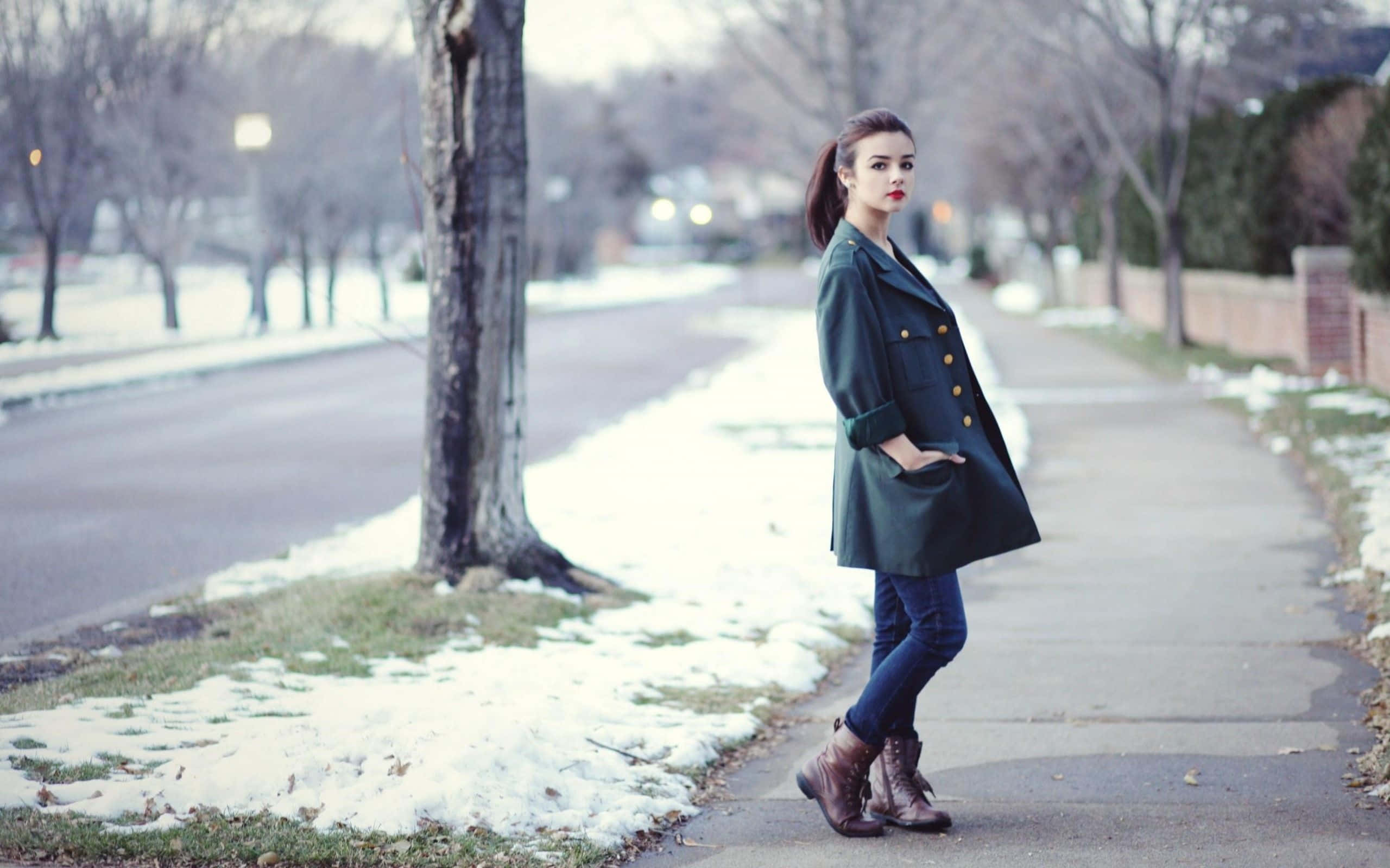 Winter Fashion 桌布 壁紙 — 一位身穿深綠色外套的女性站在寧靜冬季社區中一棵光禿禿的樹旁邊的積雪人行道上。