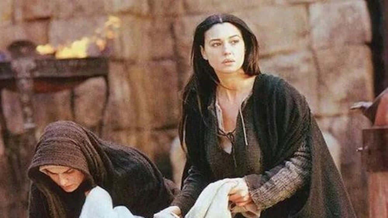Mary Magdalene 桌布 壁紙 — 一位身穿深色中世紀長袍的女性，站在另一個戴兜帽的人物旁邊，靠著石牆，火炬正在燃燒。