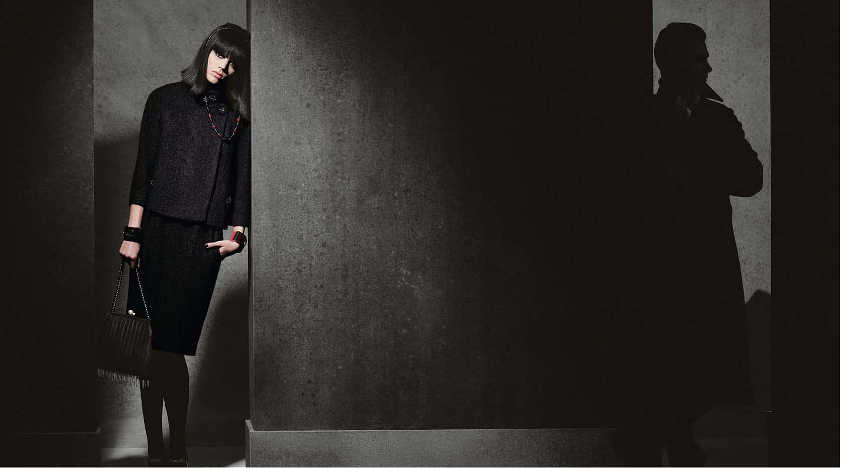 Winter Fashion 桌布 壁紙 — 一位身穿深色紋理外套的女性站在光線昏暗的走廊中，她的影子戲劇性地投射在旁邊的牆上。
