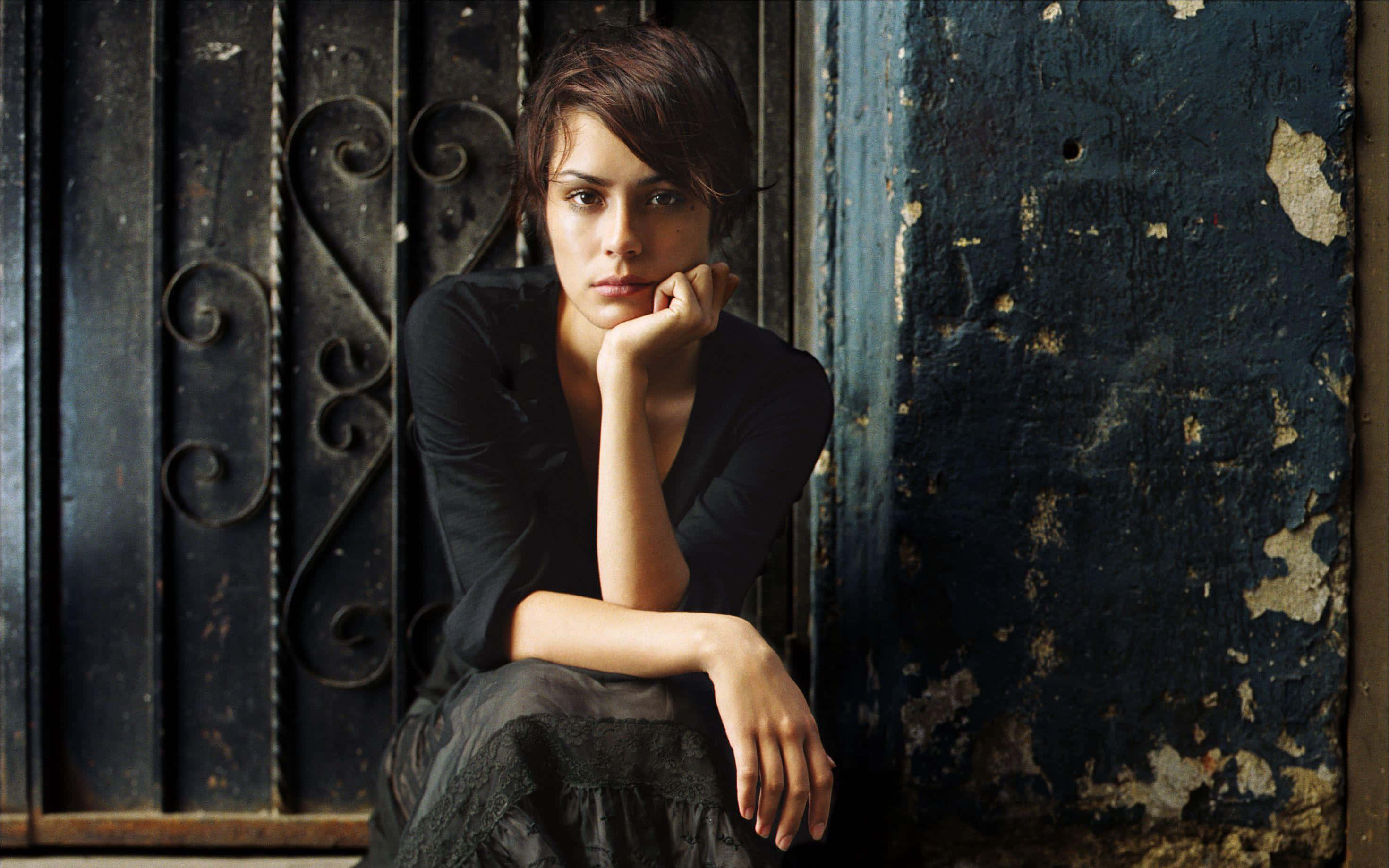 Shannyn Sossamon 桌布 壁紙 — 一位身穿深色衣服的女性靠著一扇帶有華麗金屬工藝的風化藍色門，手托下巴，若有所思地坐著。