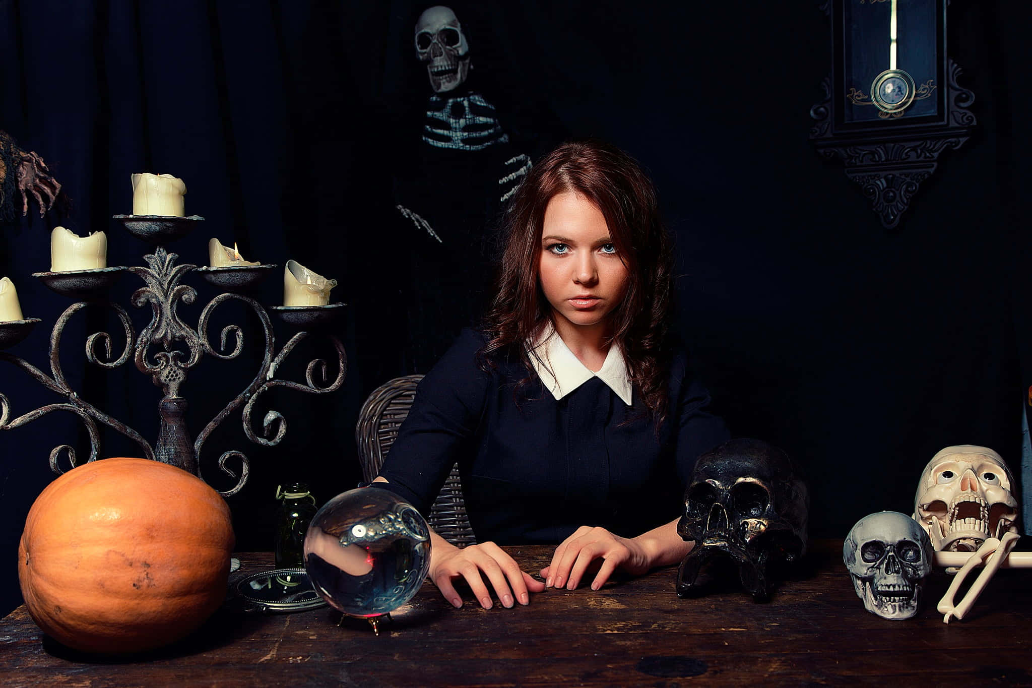 Fortune Teller 桌布 壁紙 — 一位身穿深色襯衫的年輕女性坐在木桌旁，周圍環繞著骷髏、南瓜和水晶球，燭光昏暗。