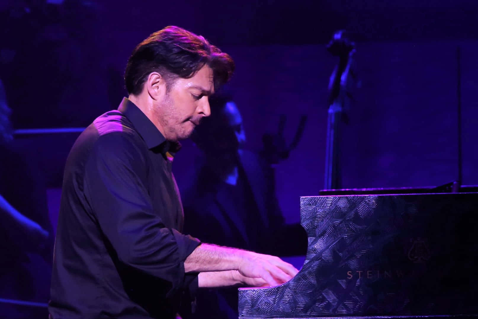 Harry Connick Jr 桌布 壁紙 — 一位身穿深色襯衫的男鋼琴家在戲劇性的紫色舞台燈光下彈奏史坦威三角鋼琴。