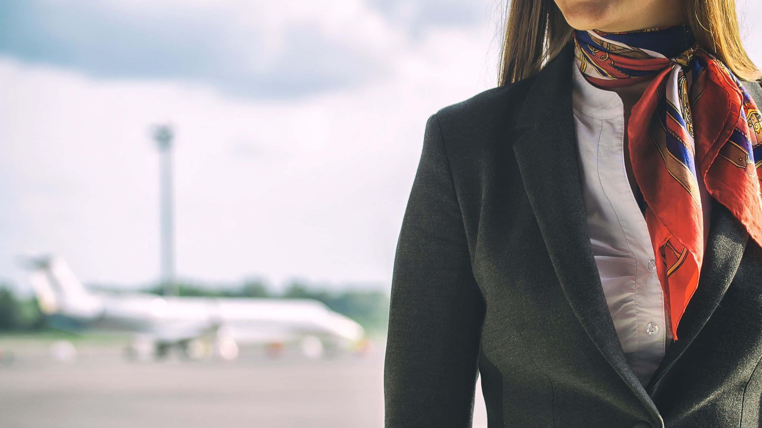 Flight Attendant 桌布 壁紙 — 一位身穿深色西裝外套和彩色圍巾的空中服務員自信地站立，背景有一架模糊的飛機。
